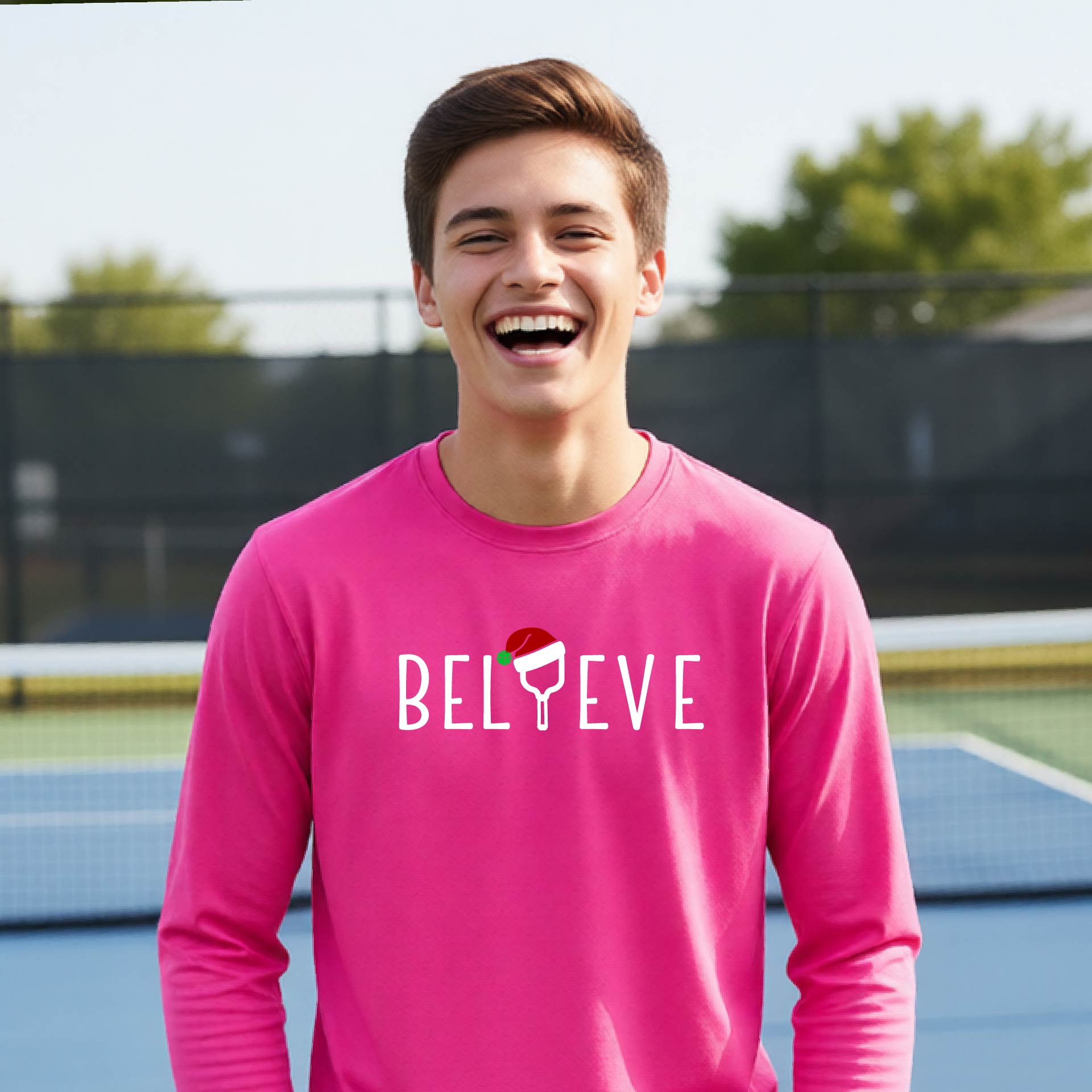 A young person laughing on a pickleball court wearing a bright pink long-sleeve shirt with 'BELIEVE' with i replaced with a pickleball paddle wearing a santa hat printed on the front.