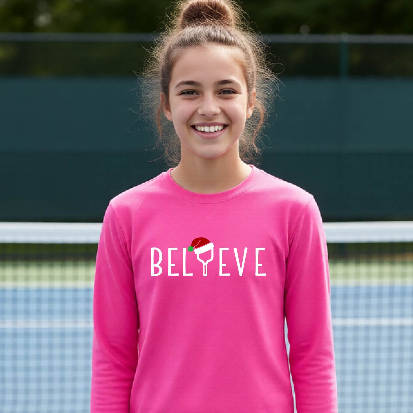 A young person laughing on a pickleball court wearing a bright pink long-sleeve shirt with 'BELIEVE' with i replaced with a pickleball paddle wearing a santa hat printed on the front.