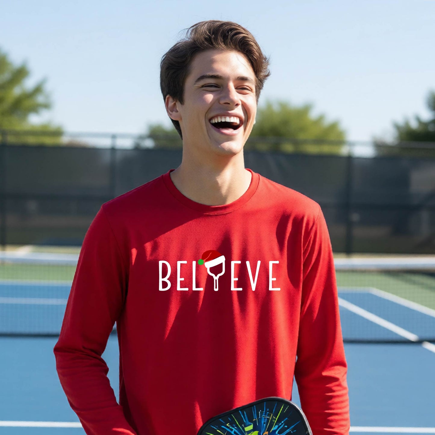 A young person laughing on a pickleball court wearing a bright red long-sleeve shirt with 'BELIEVE' with i replaced with a pickleball paddle wearing a santa hat printed on the front.