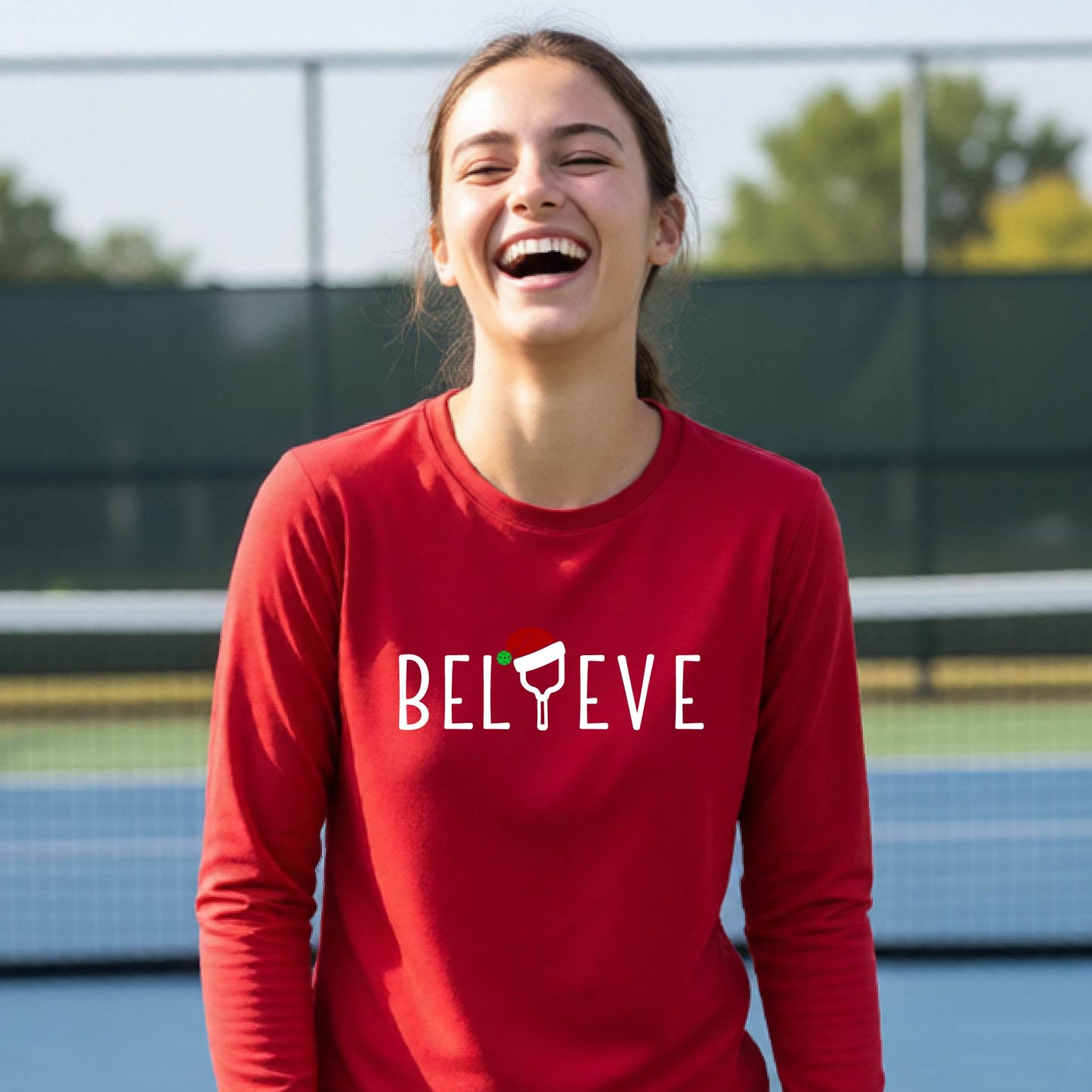 A young person laughing on a pickleball court wearing a red long-sleeve shirt with 'BELIEVE' with i replaced with a pickleball paddle wearing a santa hat printed on the front.