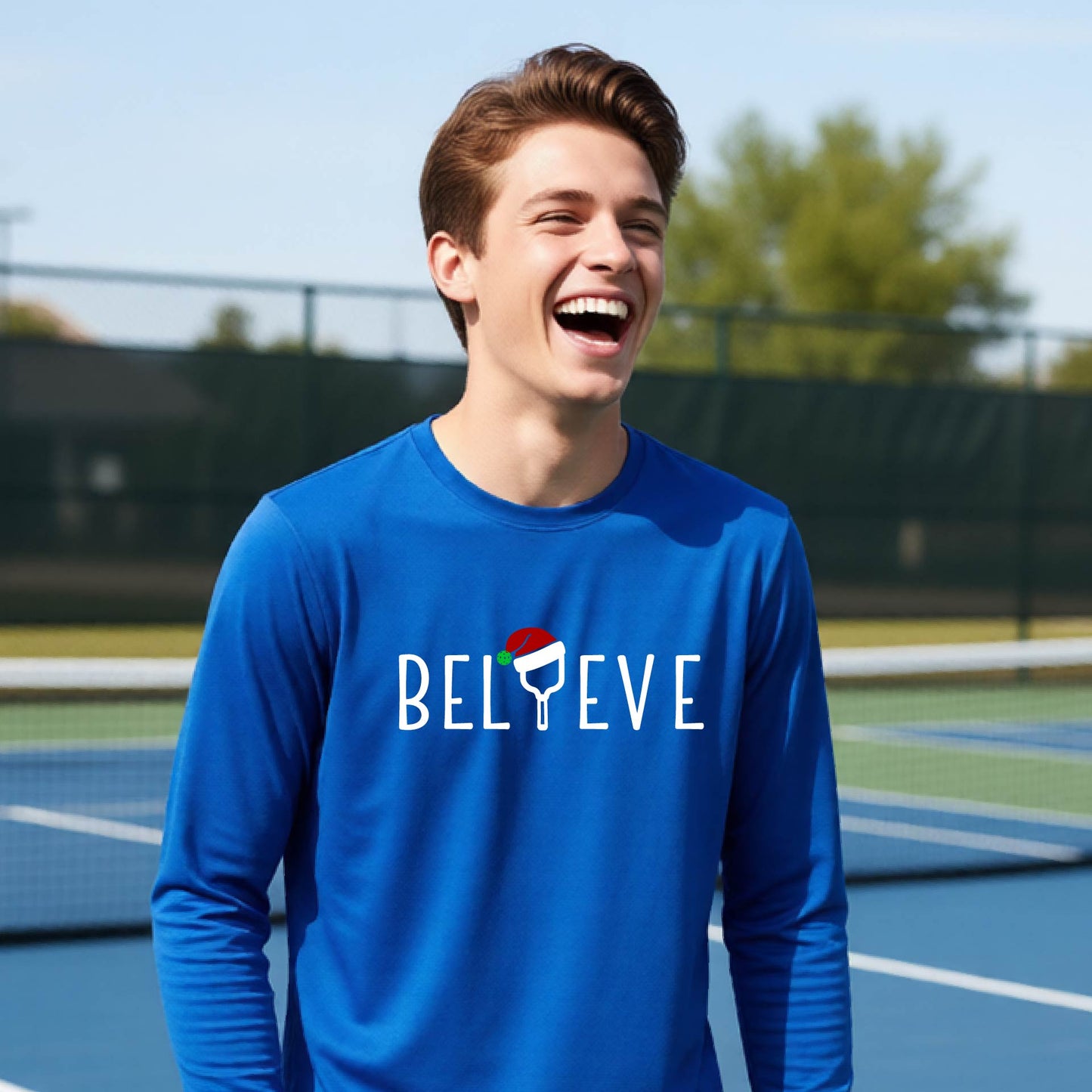 A young person laughing on a pickleball court wearing a royal blue long-sleeve shirt with 'BELIEVE' with i replaced with a pickleball paddle wearing a santa hat printed on the front.