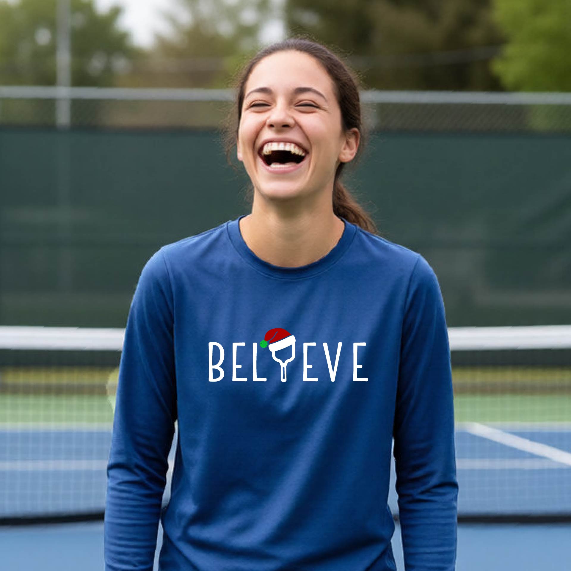 A young person laughing on a pickleball court wearing a royal blue long-sleeve shirt with 'BELIEVE' with i replaced with a pickleball paddle wearing a santa hat printed on the front.