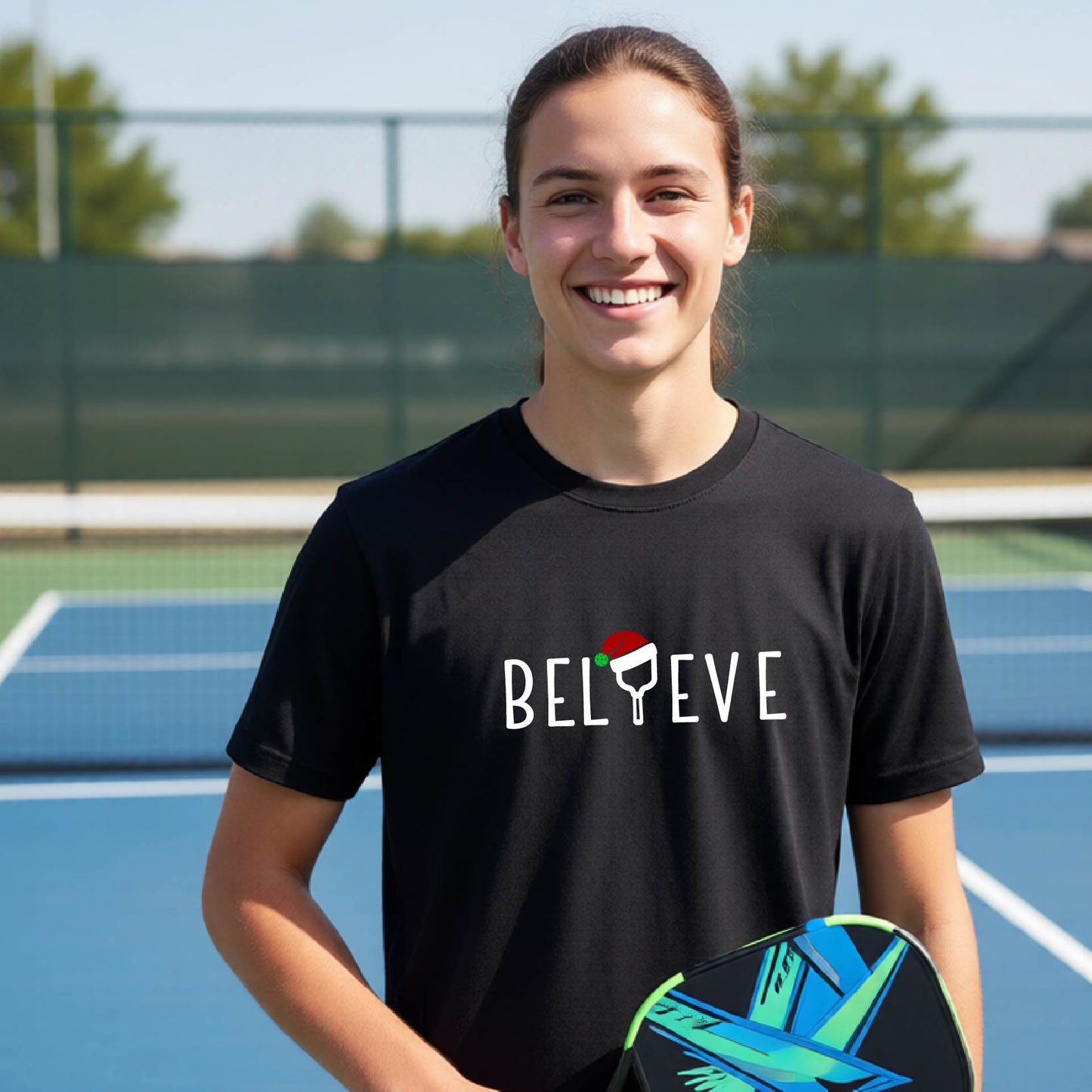 A smiling young person on a blue pickleball court wearing a black t-shirt with 'BELIEVE' printed in white, where the 'I' is replaced by a pickleball paddle topped with a red Santa hat. 