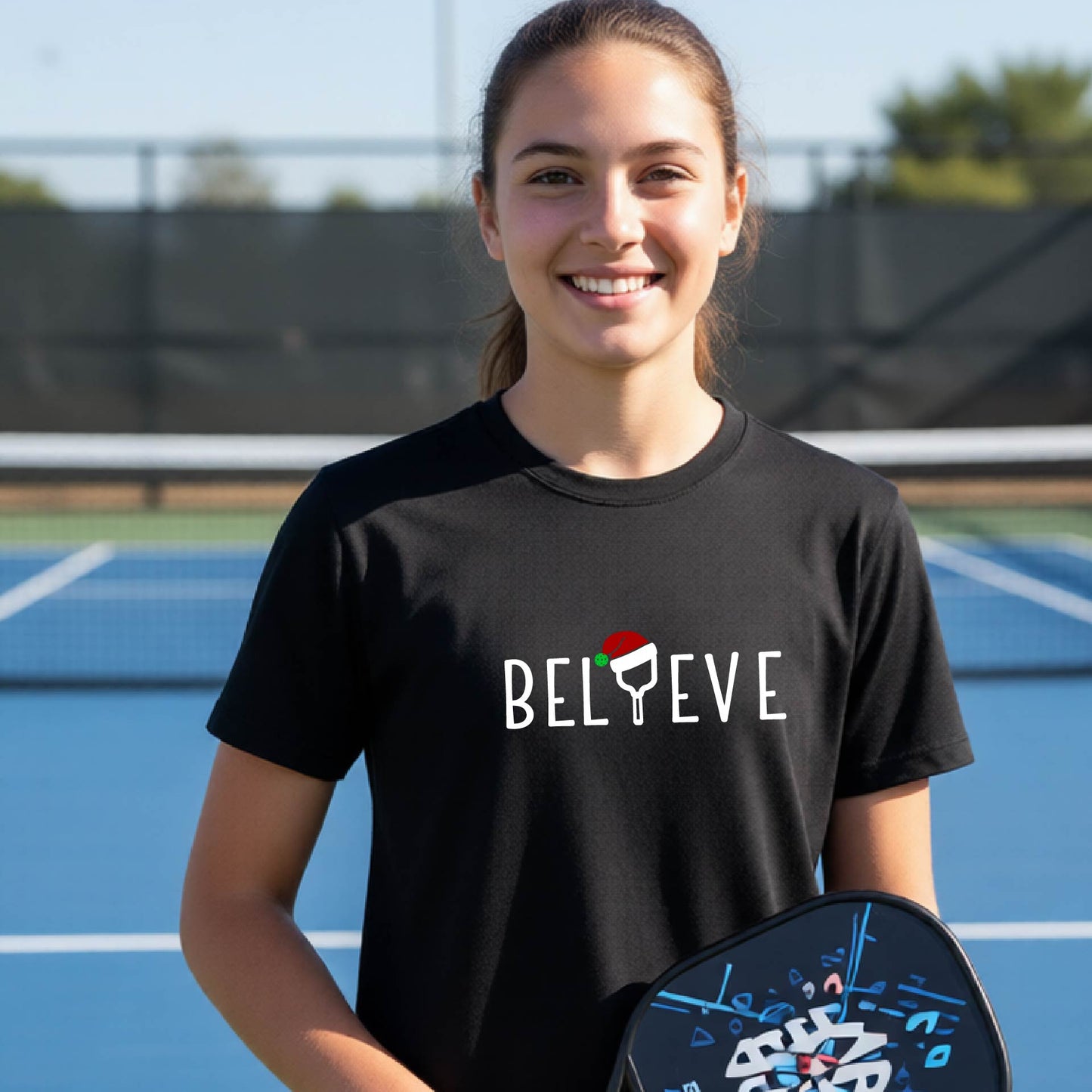 A smiling young person on a blue pickleball court wearing a black t-shirt with 'BELIEVE' printed in white, where the 'I' is replaced by a pickleball paddle topped with a red Santa hat. 