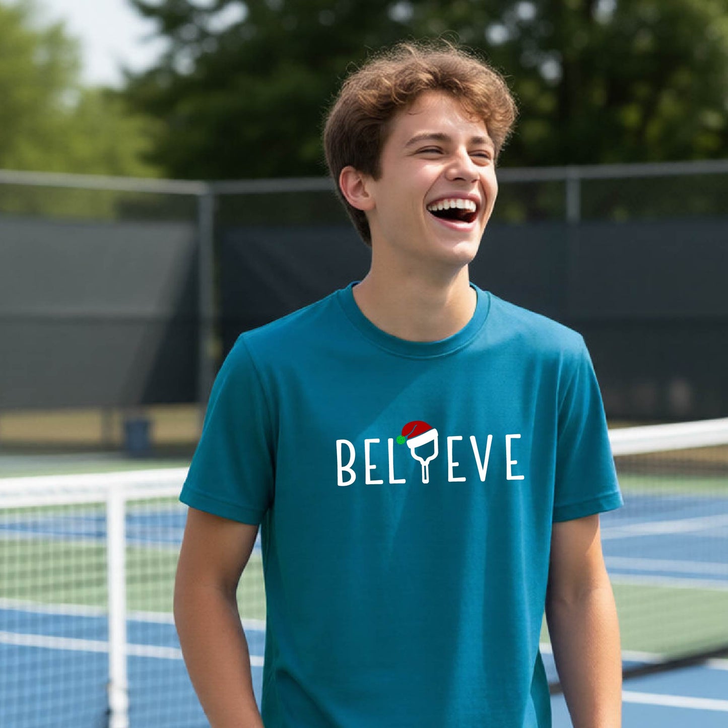 A smiling young person on a blue pickleball court wearing a teal t-shirt with 'BELIEVE' printed in white, where the 'I' is replaced by a pickleball paddle topped with a red Santa hat. 