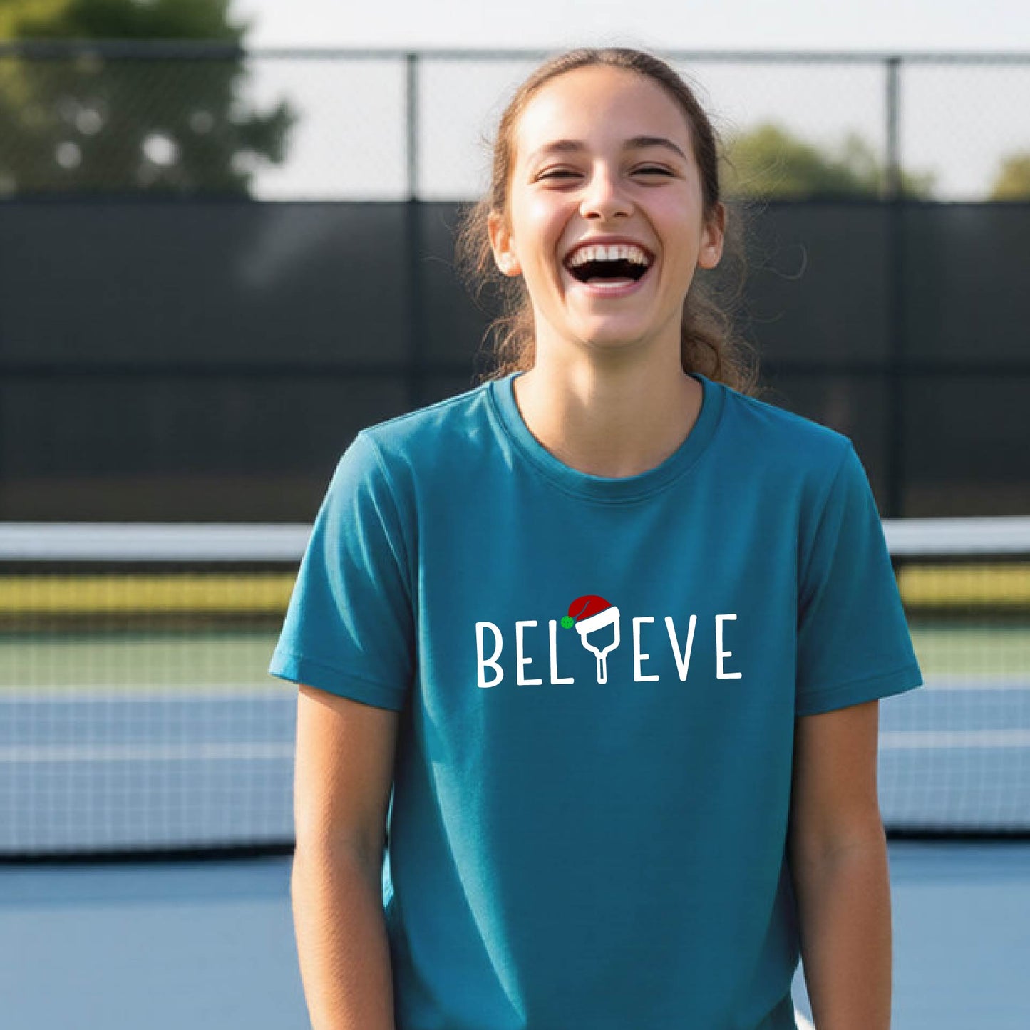 A smiling young person on a blue pickleball court wearing a teal t-shirt with 'BELIEVE' printed in white, where the 'I' is replaced by a pickleball paddle topped with a red Santa hat. 
