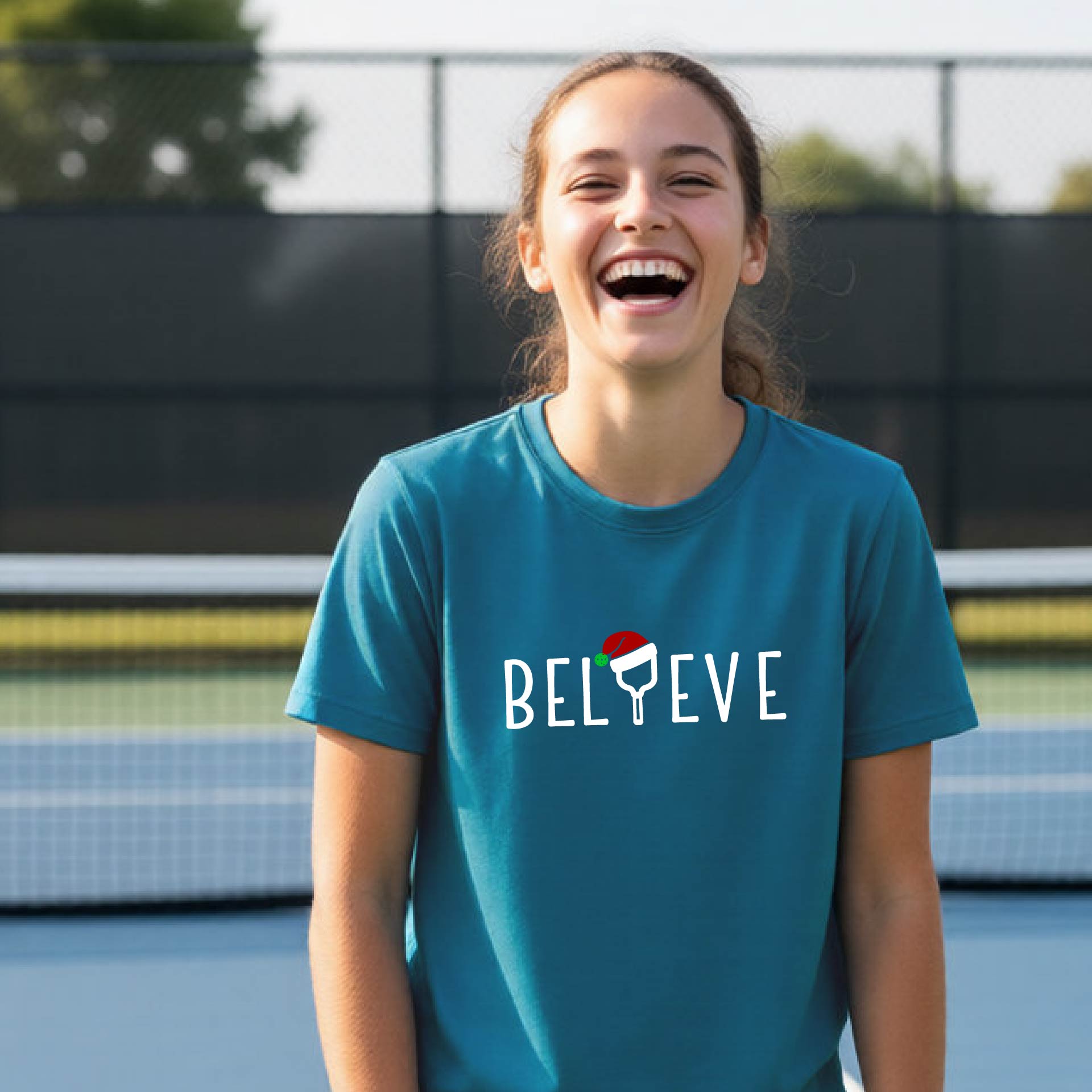 A smiling young person on a blue pickleball court wearing a teal t-shirt with 'BELIEVE' printed in white, where the 'I' is replaced by a pickleball paddle topped with a red Santa hat. 