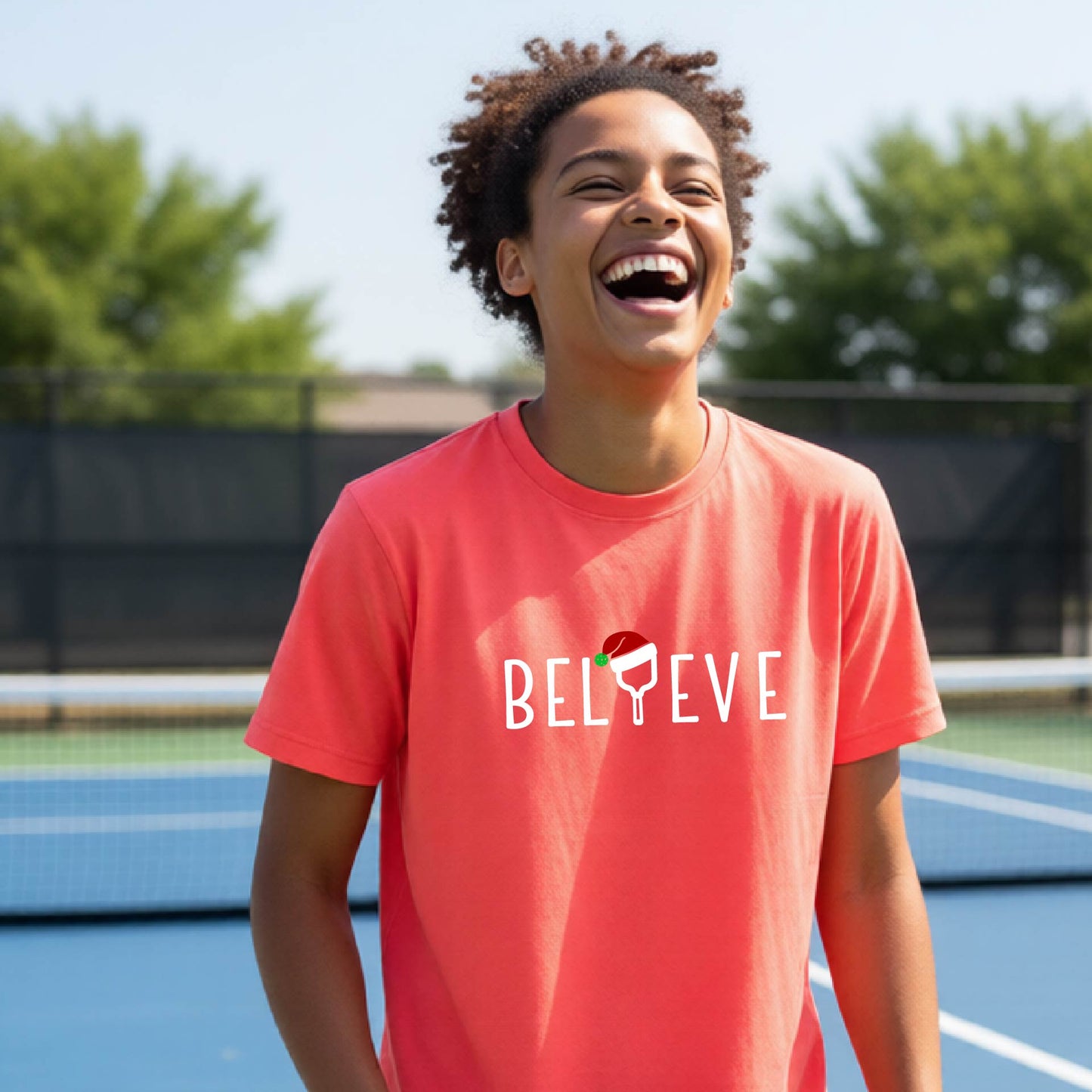 A smiling young person on a blue pickleball court wearing a coral-colored t-shirt with 'BELIEVE' printed in white, where the 'I' is replaced by a pickleball paddle topped with a red Santa hat. 