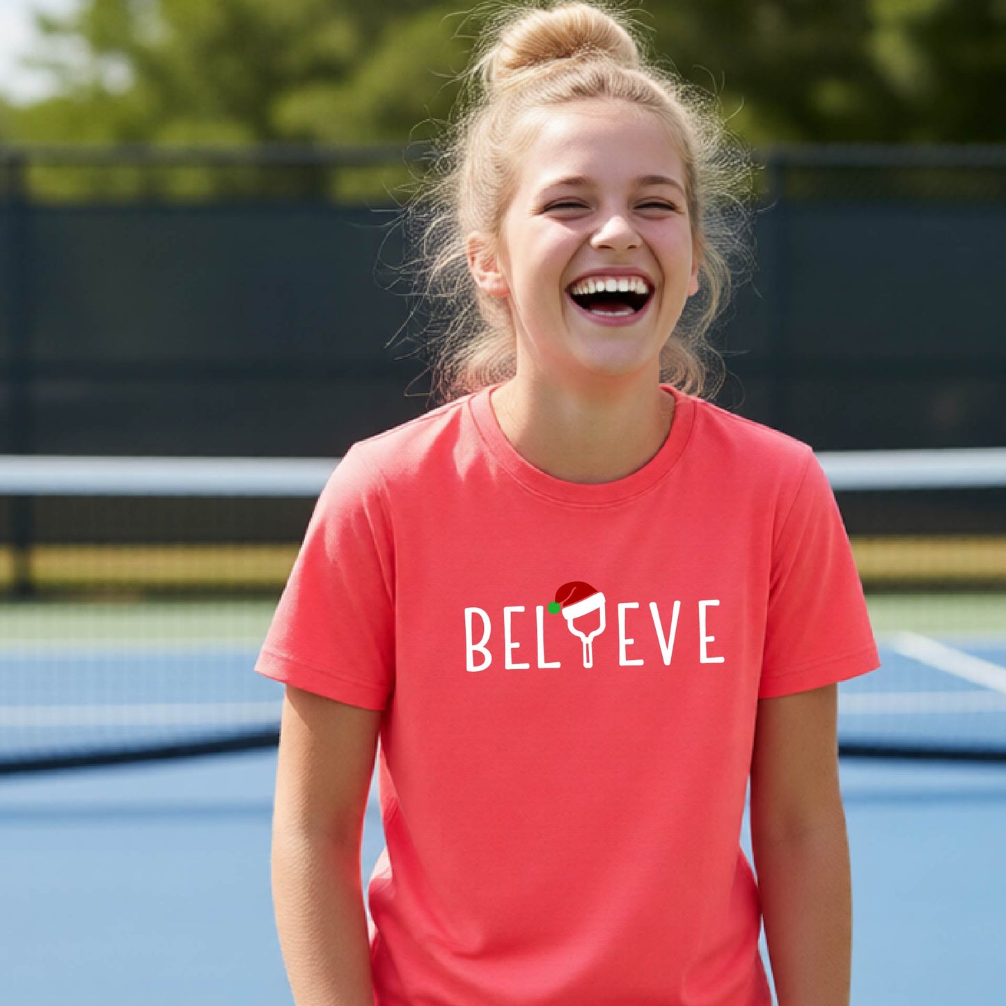A smiling young person on a blue pickleball court wearing a coral-colored t-shirt with 'BELIEVE' printed in white, where the 'I' is replaced by a pickleball paddle topped with a red Santa hat. 