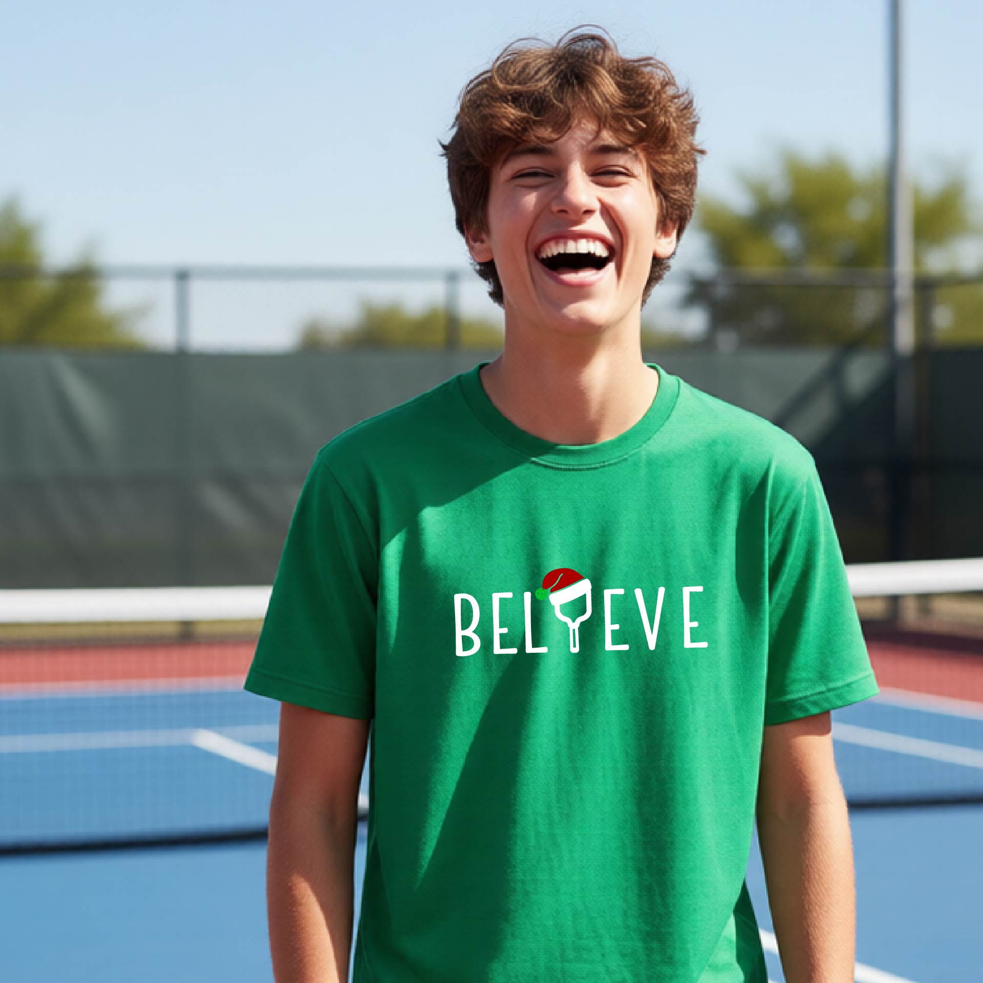 A smiling young person on a blue pickleball court wearing a green t-shirt with 'BELIEVE' printed in white, where the 'I' is replaced by a pickleball paddle topped with a red Santa hat. 