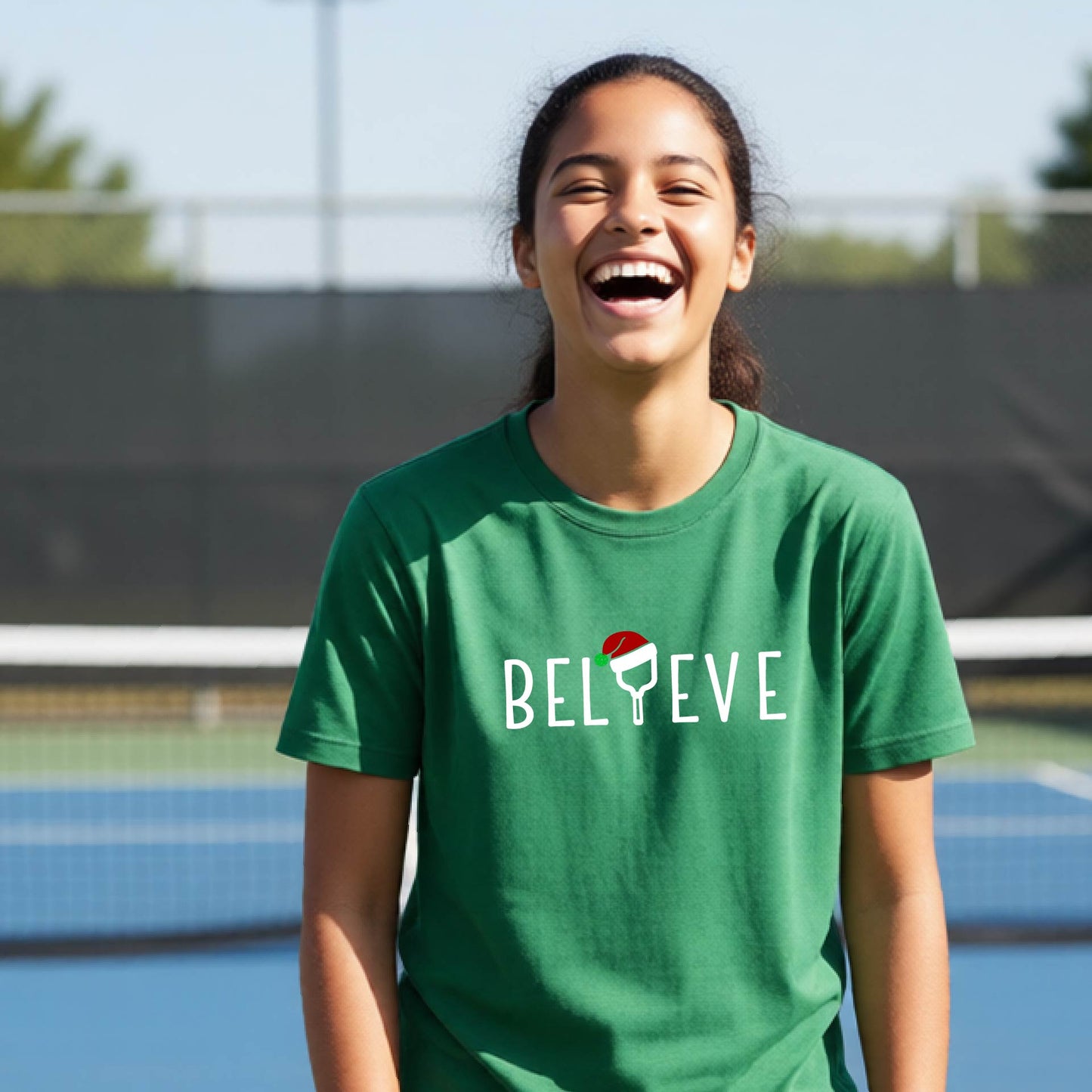 A smiling young person on a blue pickleball court wearing a green t-shirt with 'BELIEVE' printed in white, where the 'I' is replaced by a pickleball paddle topped with a red Santa hat. 