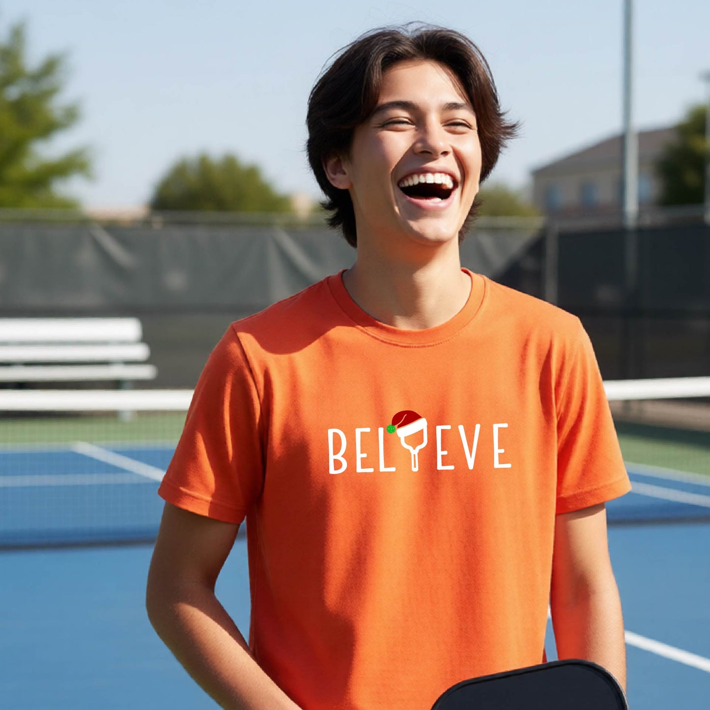 A smiling young person on a blue pickleball court wearing an orange t-shirt with 'BELIEVE' printed in white, where the 'I' is replaced by a pickleball paddle topped with a red Santa hat. 