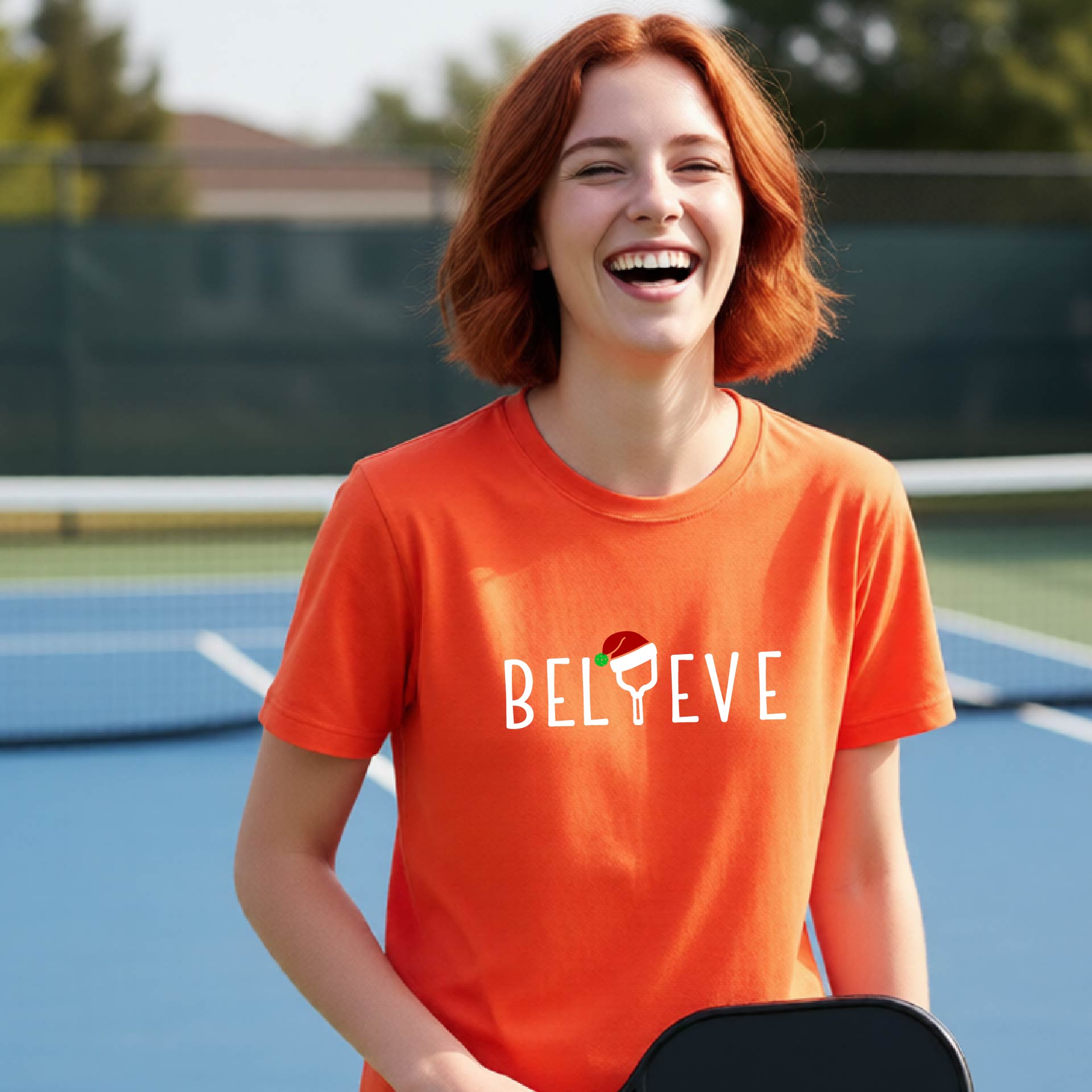 A smiling young person on a blue pickleball court wearing an orange t-shirt with 'BELIEVE' printed in white, where the 'I' is replaced by a pickleball paddle topped with a red Santa hat. 