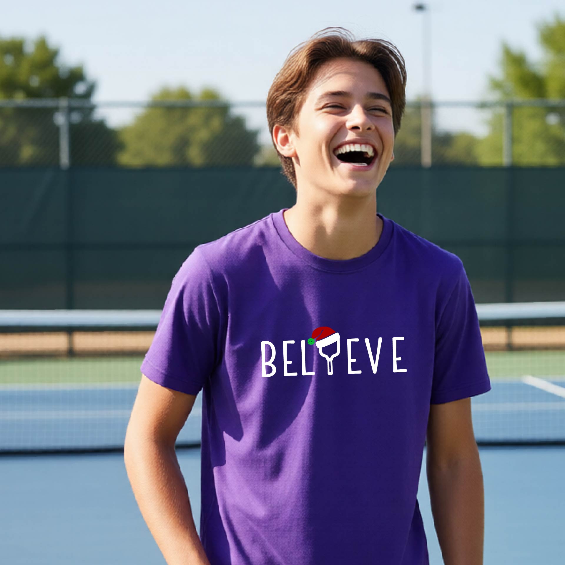 A smiling young person on a blue pickleball court wearing a purple t-shirt with 'BELIEVE' printed in white, where the 'I' is replaced by a pickleball paddle topped with a red Santa hat. 