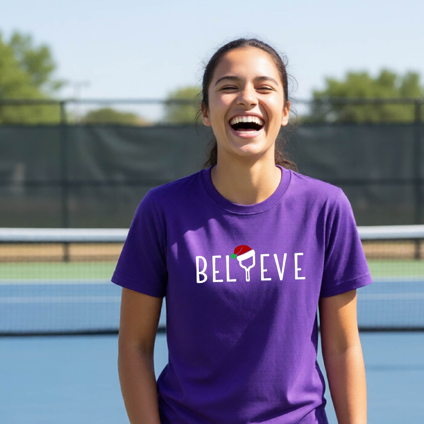 A smiling young person on a blue pickleball court wearing a purple t-shirt with 'BELIEVE' printed in white, where the 'I' is replaced by a pickleball paddle topped with a red Santa hat. 
