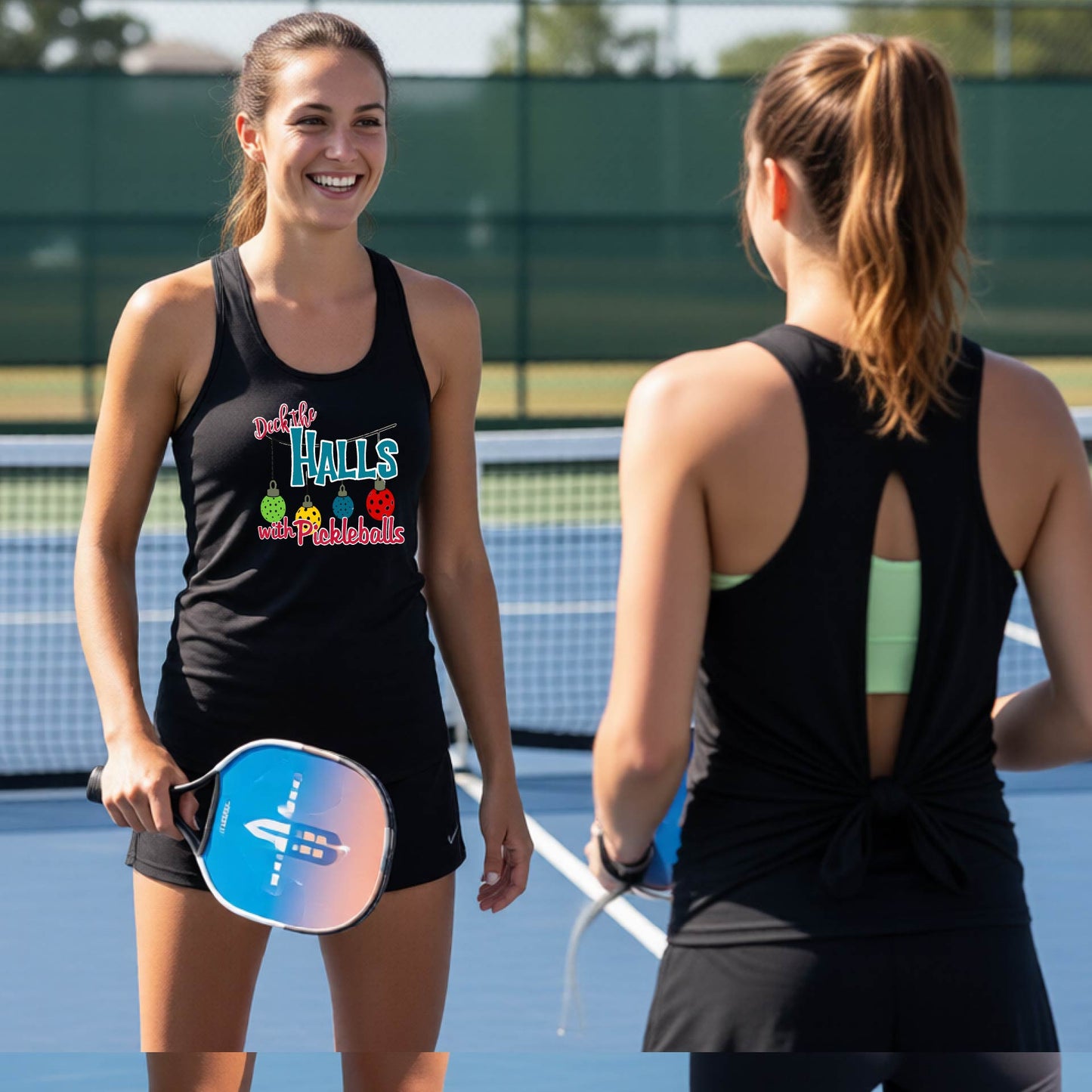 Two women on a pickleball court wearing matching black athletic tank tops. The woman facing forward has a pickleball graphic with the words "DECK THA HALLS" onthe front. 