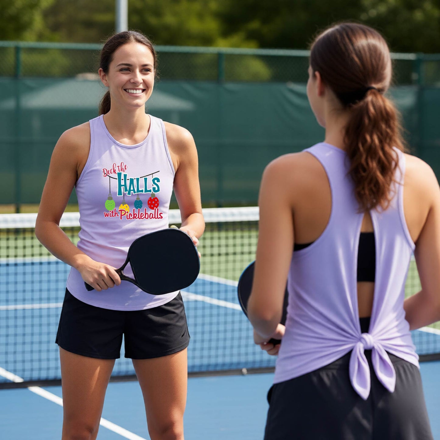 Two women on a pickleball court wearing matching light lavender athletic tank tops. The woman facing forward has a pickleball graphic with the words "DECK THA HALLS" onthe front. 