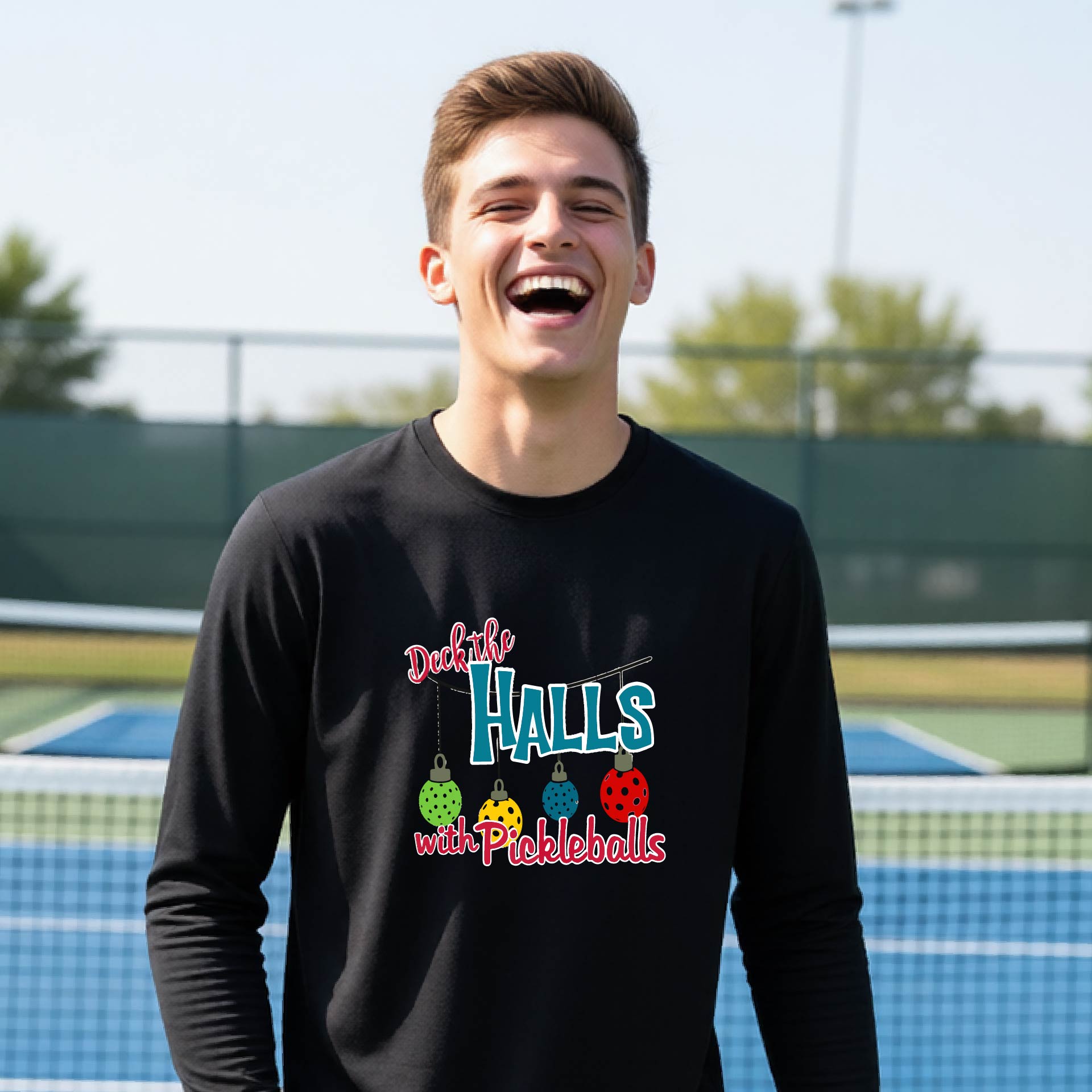 A smiling young person on a pickleball court wearing a black long-sleeve graphic shirt that reads 'Deck the Halls with Pickleballs' .