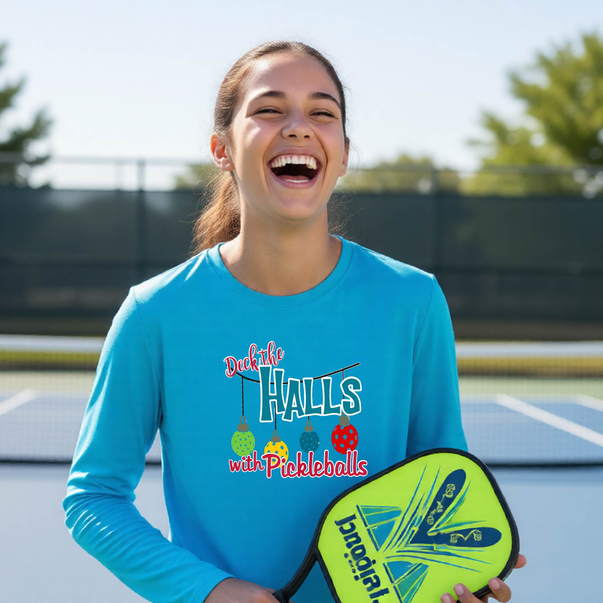 A smiling young person on a pickleball court wearing a bright blue long-sleeve graphic shirt that reads 'Deck the Halls with Pickleballs' .
