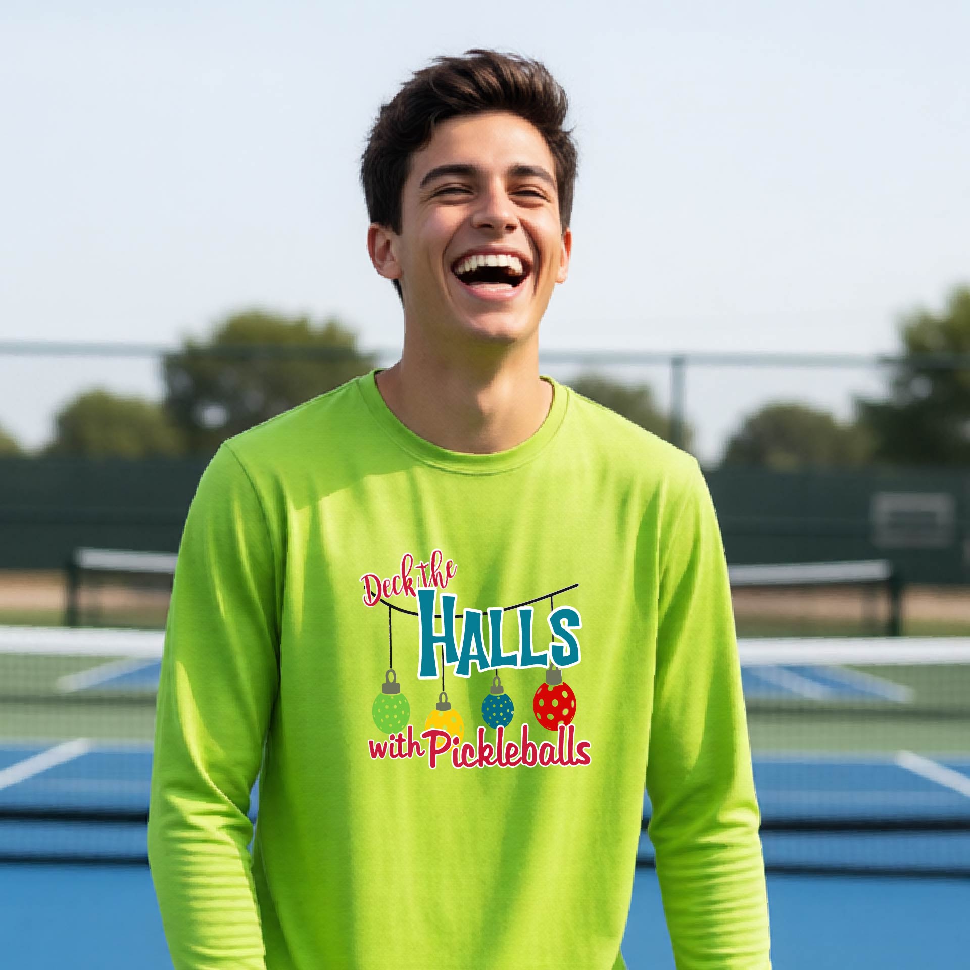 A smiling young person on a pickleball court wearing a bright lime green long-sleeve graphic shirt that reads 'Deck the Halls with Pickleballs' .