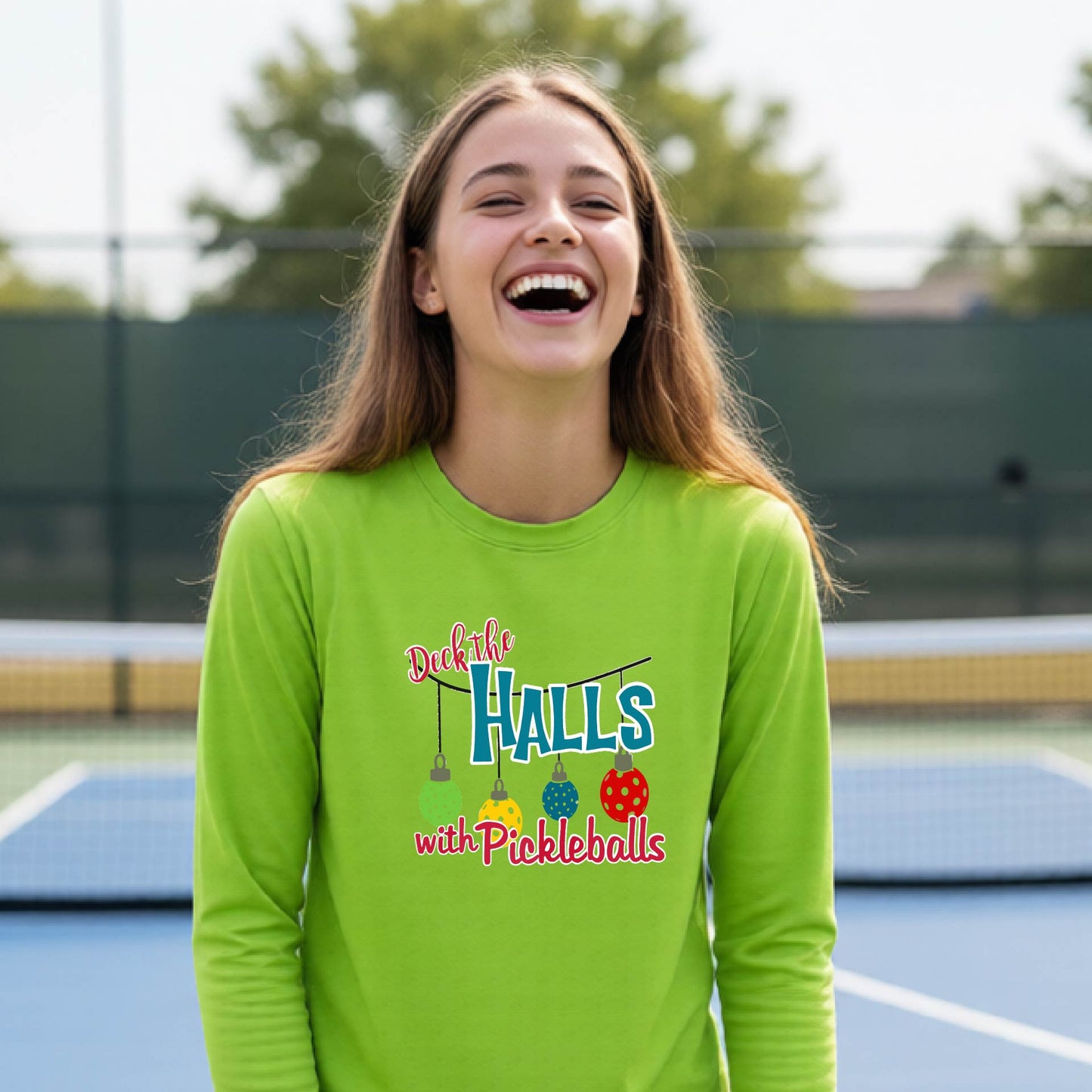 A smiling young person on a pickleball court wearing a bright lime green long-sleeve graphic shirt that reads 'Deck the Halls with Pickleballs' .