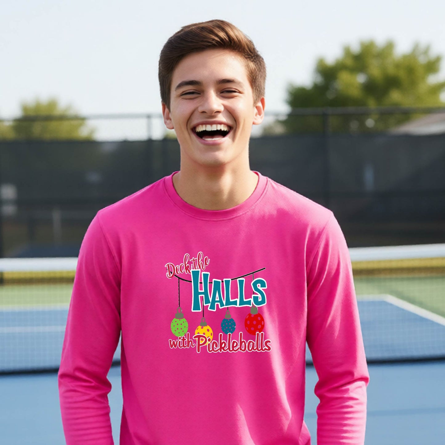 A smiling young person on a pickleball court wearing a bright pink long-sleeve graphic shirt that reads 'Deck the Halls with Pickleballs' .