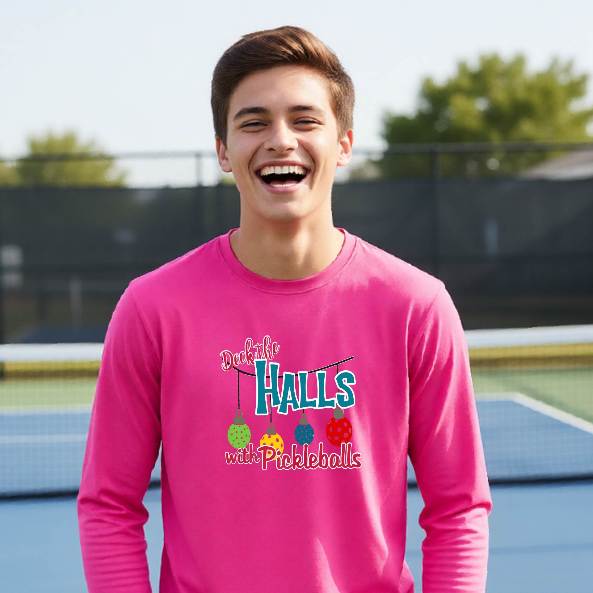 A smiling young person on a pickleball court wearing a bright pink long-sleeve graphic shirt that reads 'Deck the Halls with Pickleballs' .