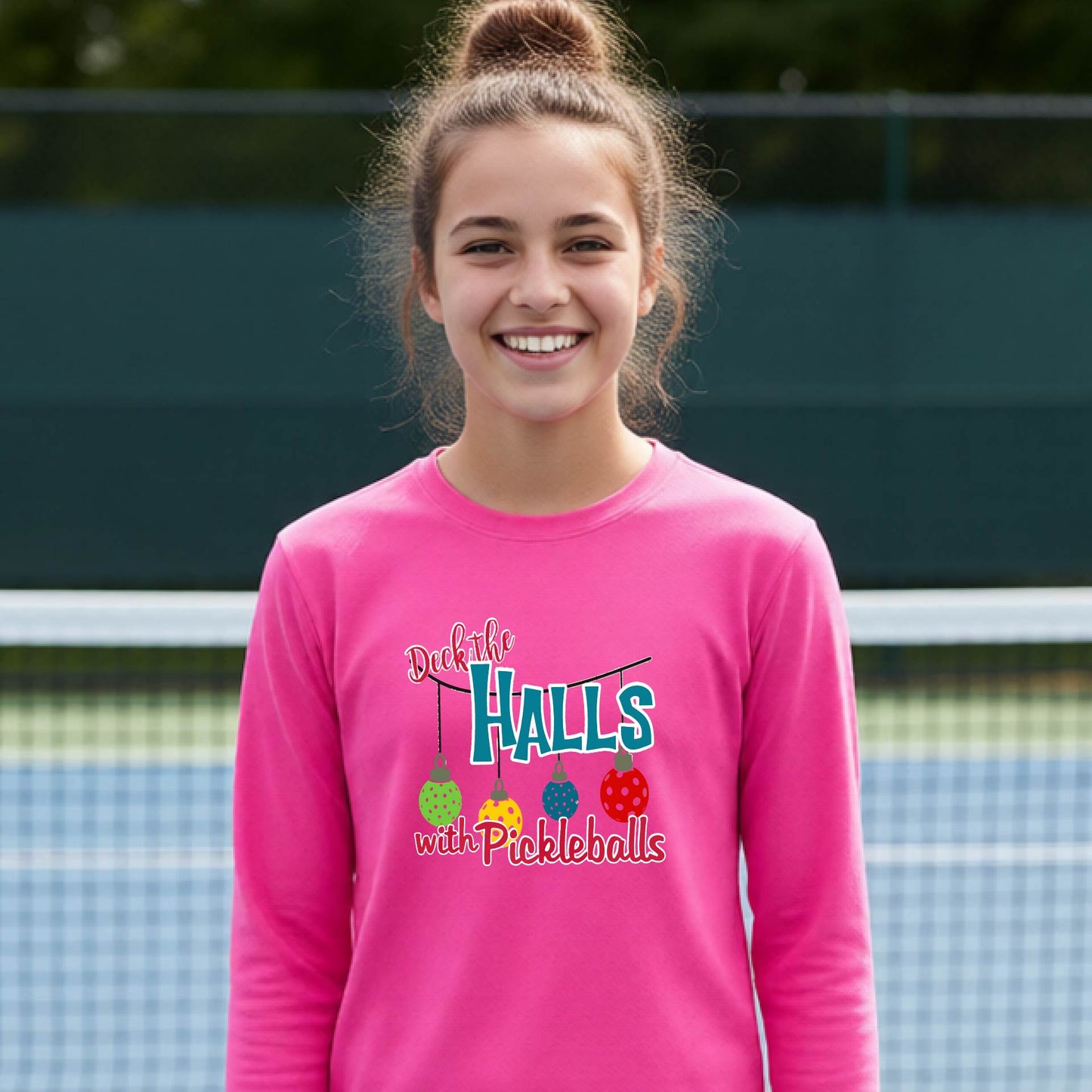 A smiling young person on a pickleball court wearing a bright pink long-sleeve graphic shirt that reads 'Deck the Halls with Pickleballs' .