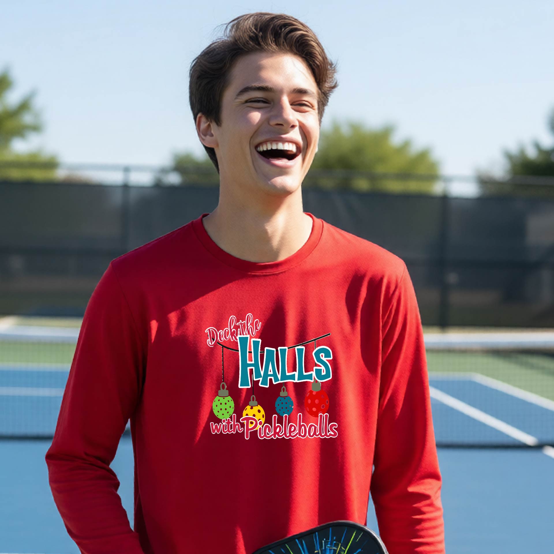 A smiling young person on a pickleball court wearing a bright red long-sleeve graphic shirt that reads 'Deck the Halls with Pickleballs' .