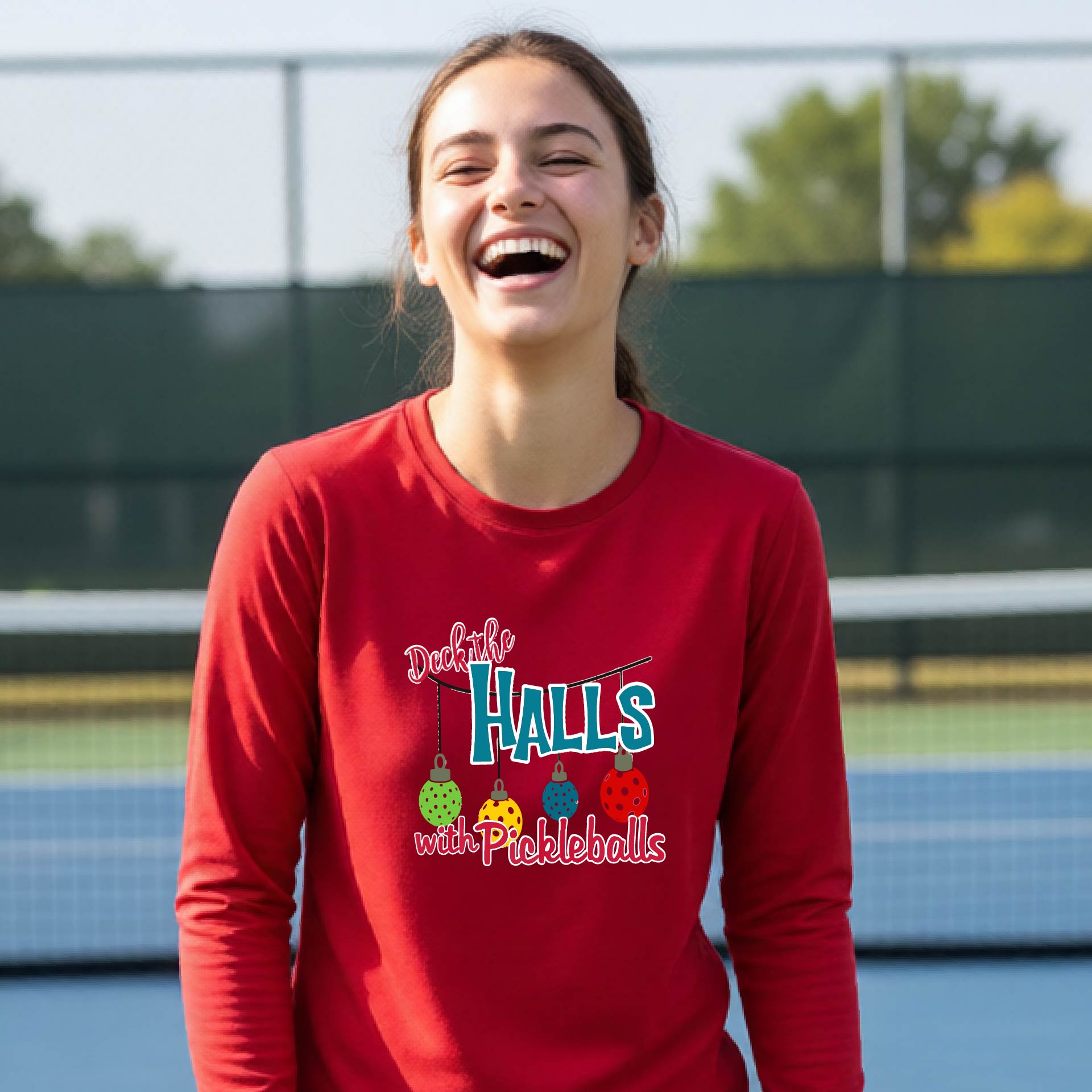 A smiling young person on a pickleball court wearing a bright red long-sleeve graphic shirt that reads 'Deck the Halls with Pickleballs' .