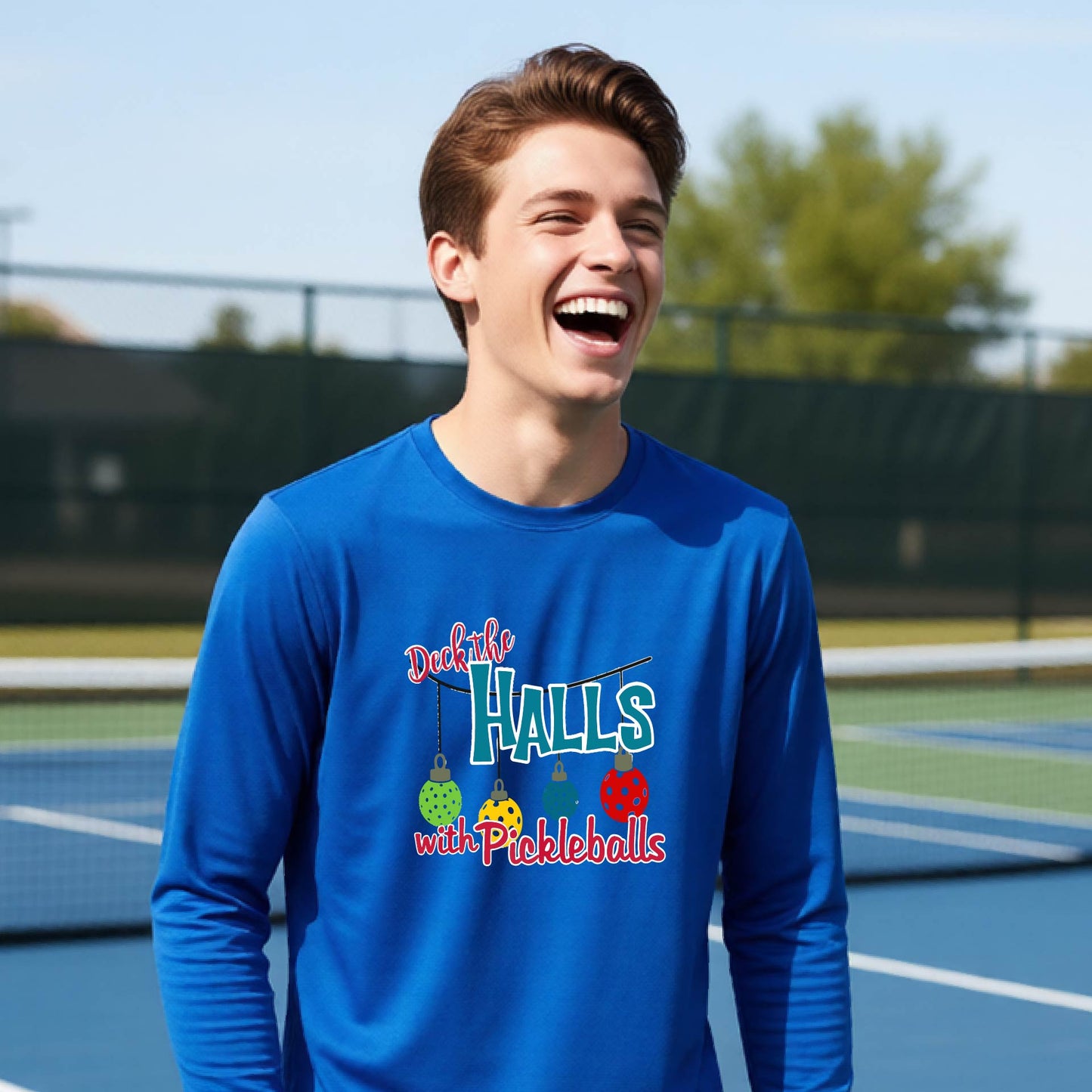 A smiling young person on a pickleball court wearing a royal  blue long-sleeve graphic shirt that reads 'Deck the Halls with Pickleballs' .