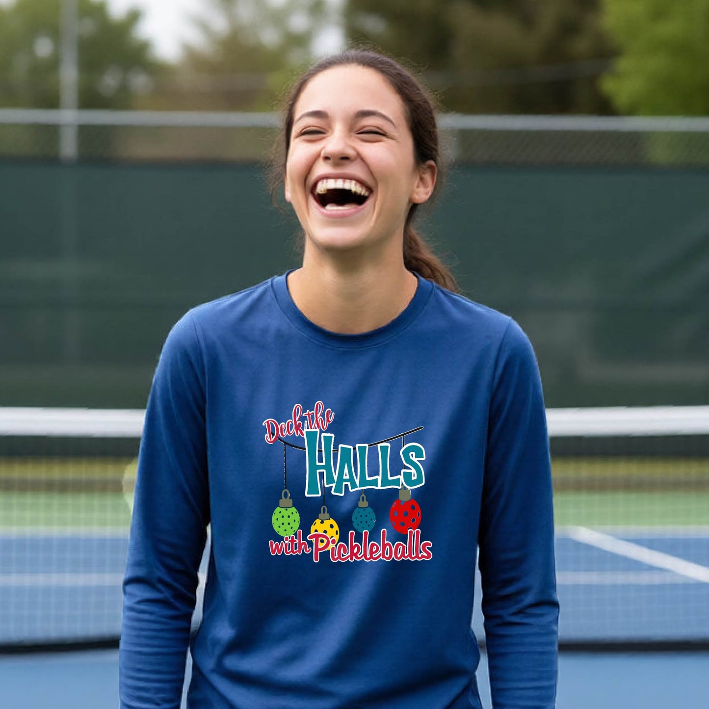 A smiling young person on a pickleball court wearing a royal blue long-sleeve graphic shirt that reads 'Deck the Halls with Pickleballs' .