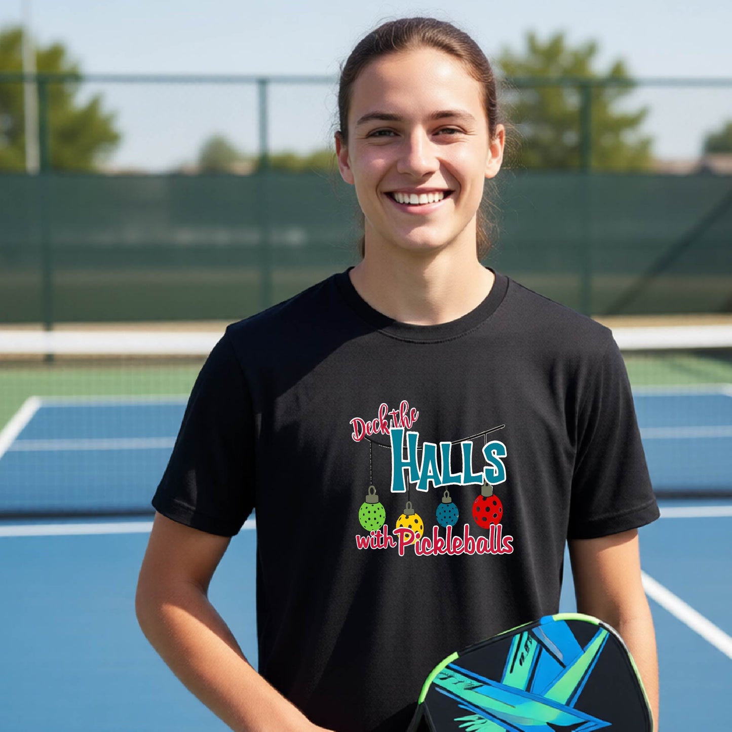 A smiling young woman stands on a blue pickleball court wearing a black graphic t-shirt that reads 'Deck the HALLS with Pickleballs' in colorful lettering, holding a pickleball paddle.