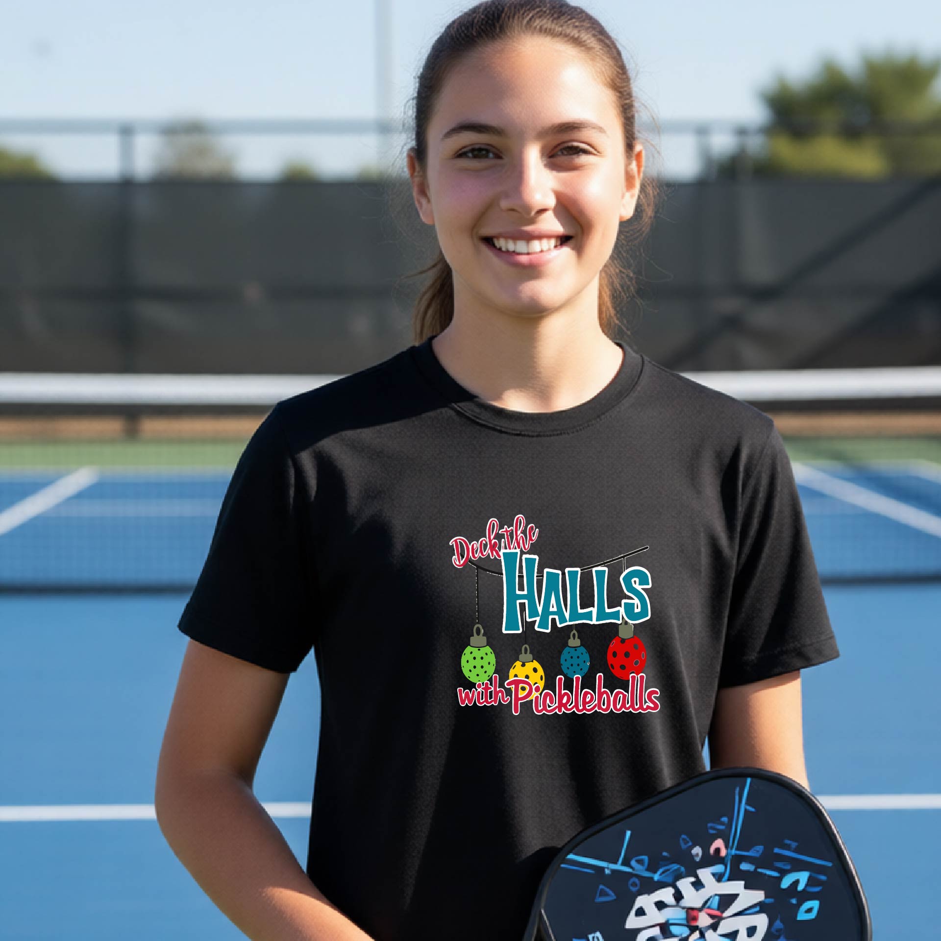 A smiling young woman stands on a blue pickleball court wearing a black graphic t-shirt that reads 'Deck the Halls with Pickleballs' with colorful pickleballs and a pickleball paddle in her hand.