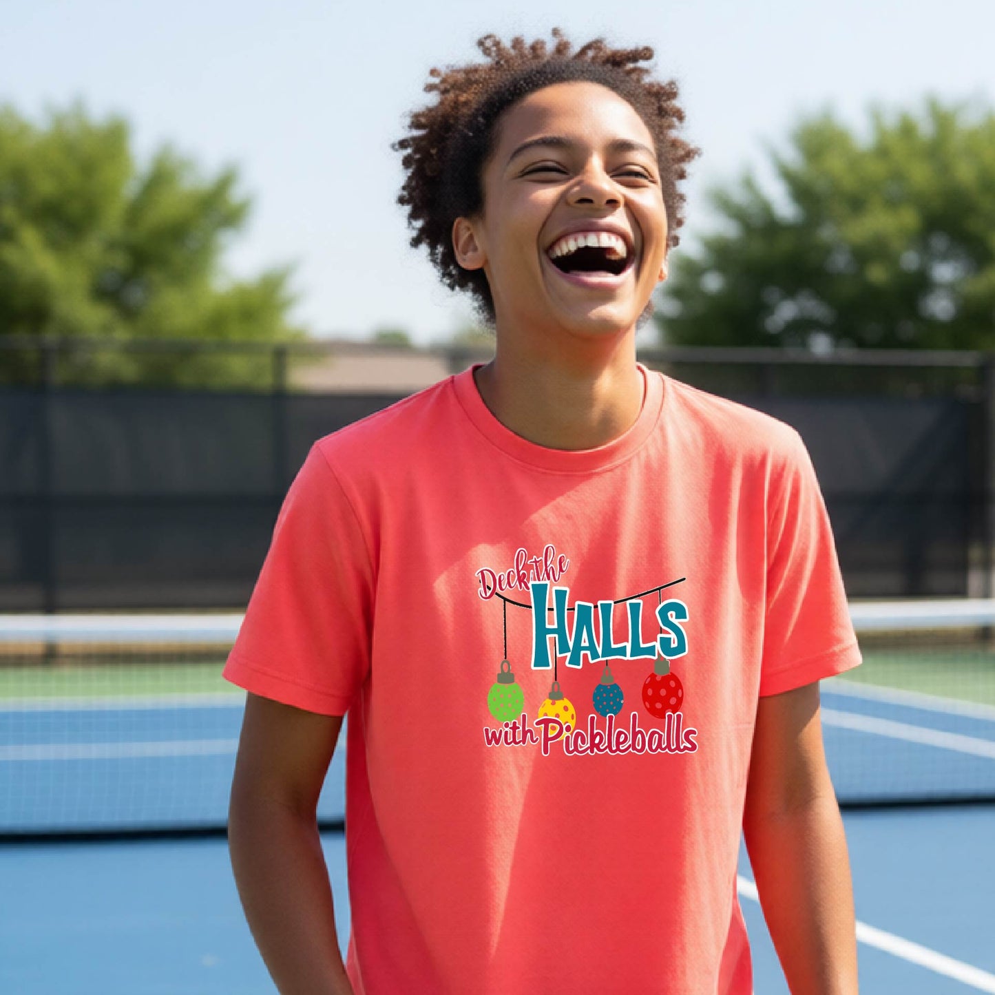 A young person laughing on an outdoor pickleball court wearing a coral-pink t-shirt with a colorful graphic that reads 'Deck the Halls with Pickleballs'.