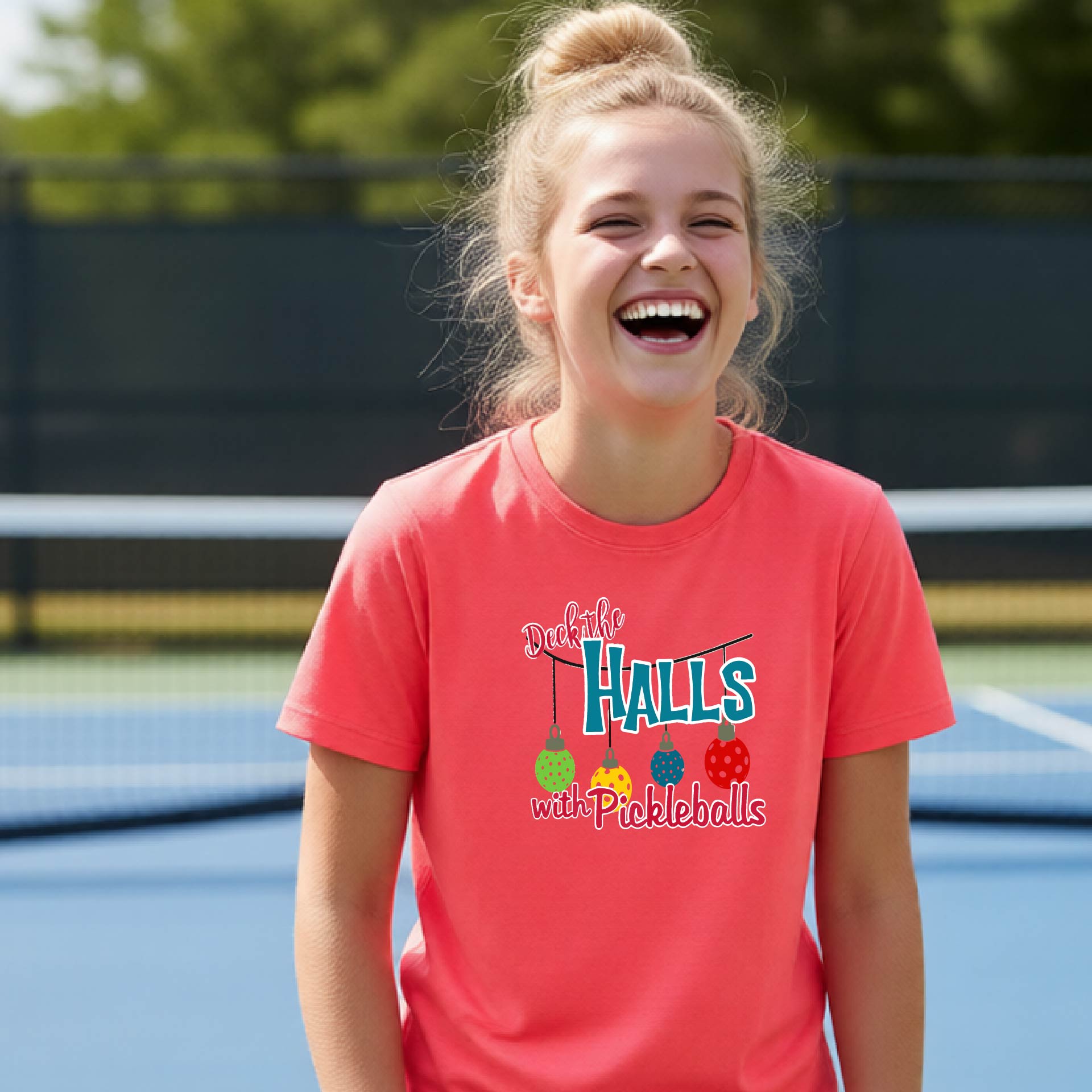 A young girl with blonde hair in a bun is laughing on an outdoor pickleball court, wearing a coral pink t-shirt with a graphic that reads 'Deck the HALLS with Pickleballs' and features colorful pickleballs.