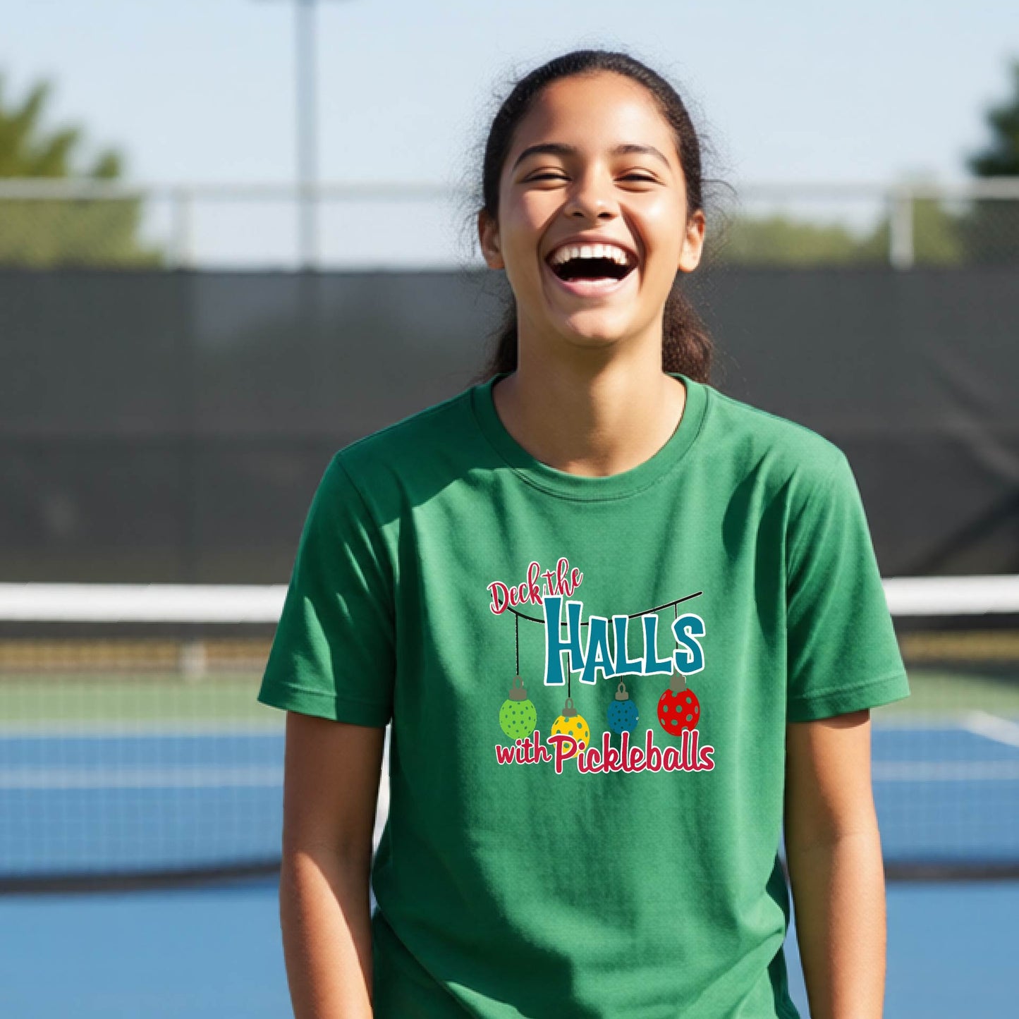 A young woman smiling on an outdoor pickleball court wearing a green graphic t-shirt that reads 'Deck the HALLS with Pickleballs' in blue, pink, and yellow lettering.