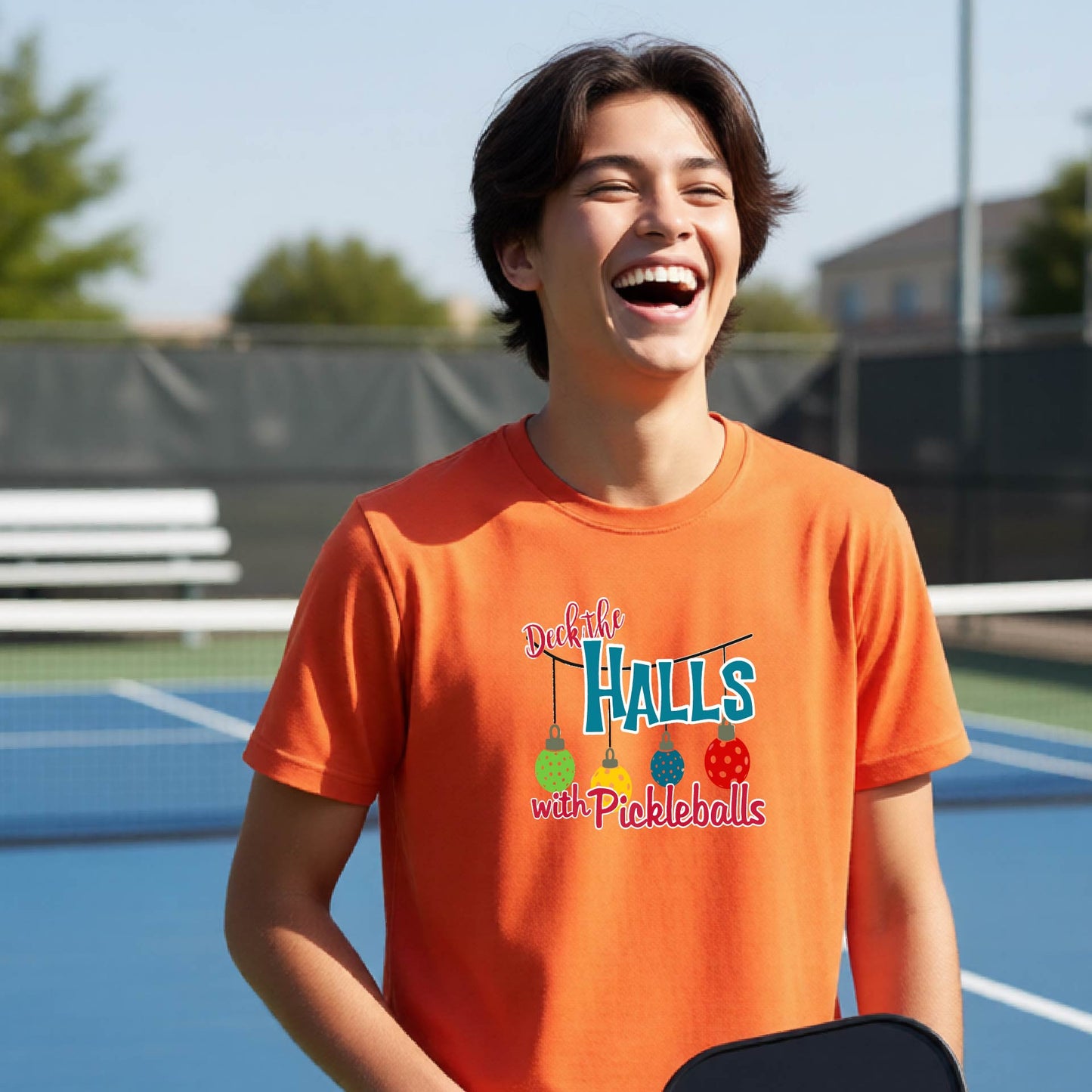 A young man laughing on an outdoor pickleball court wearing a bright orange t-shirt with a colorful graphic that reads 'Deck the  HALLS with Pickleballs'.