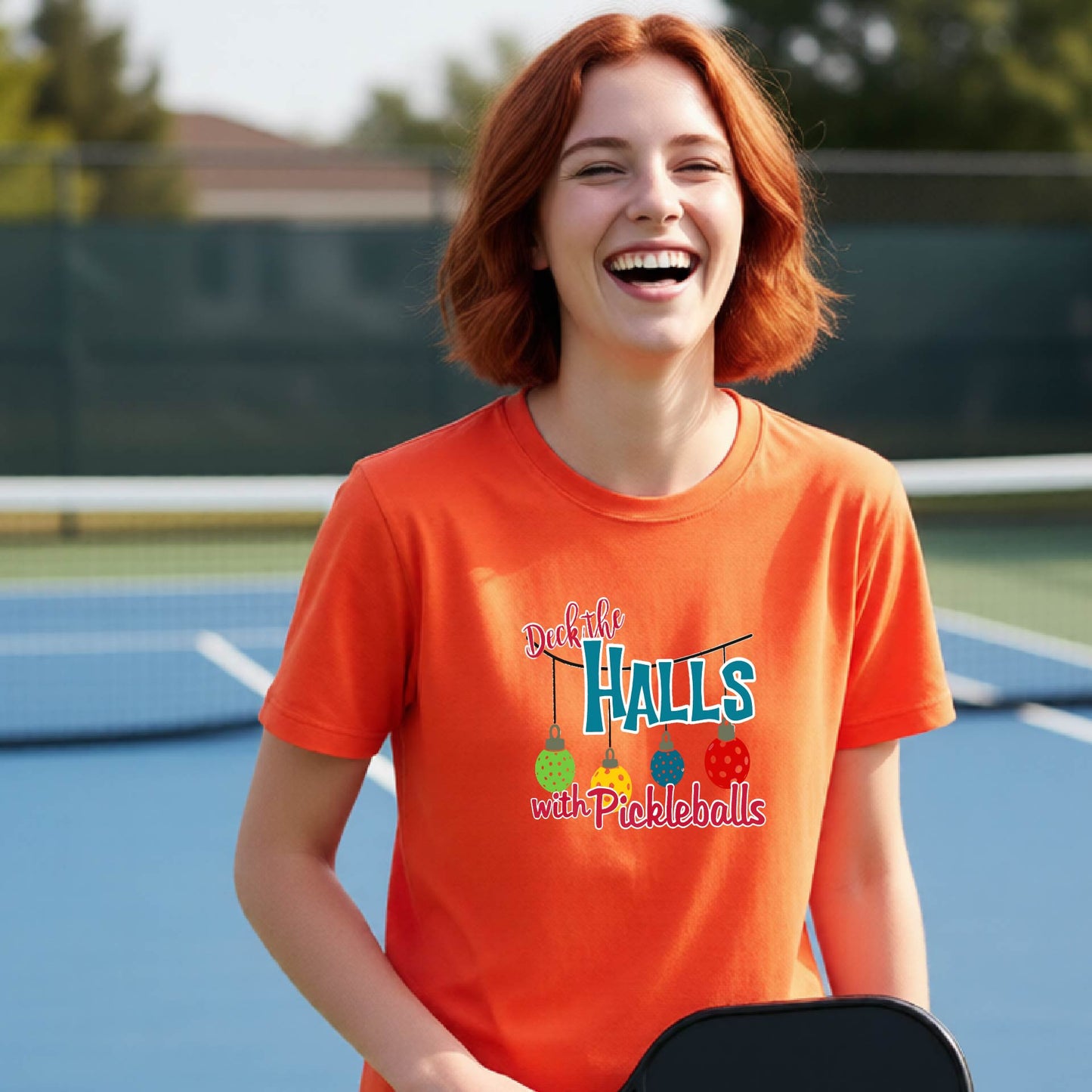 A smiling young woman with red hair stands on a blue pickleball court wearing a bright orange graphic T-shirt that reads "Deck the HALLS with Pickleballs" with colorful hanging ball illustrations. No visible brand logo is present.