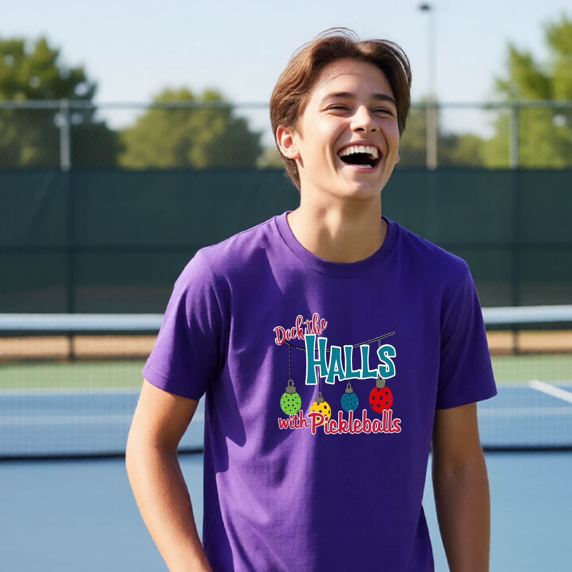 A young man laughing on an outdoor pickleball court wearing a purple t-shirt with a colorful graphic that reads 'Dck the Hall's with Pickleballs'.