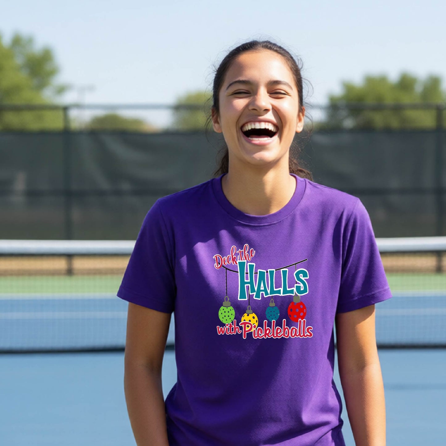 A woman laughing on an outdoor pickleball court wearing a purple t-shirt with a colorful graphic that reads 'Deck the  HALLS with Pickleballs'.