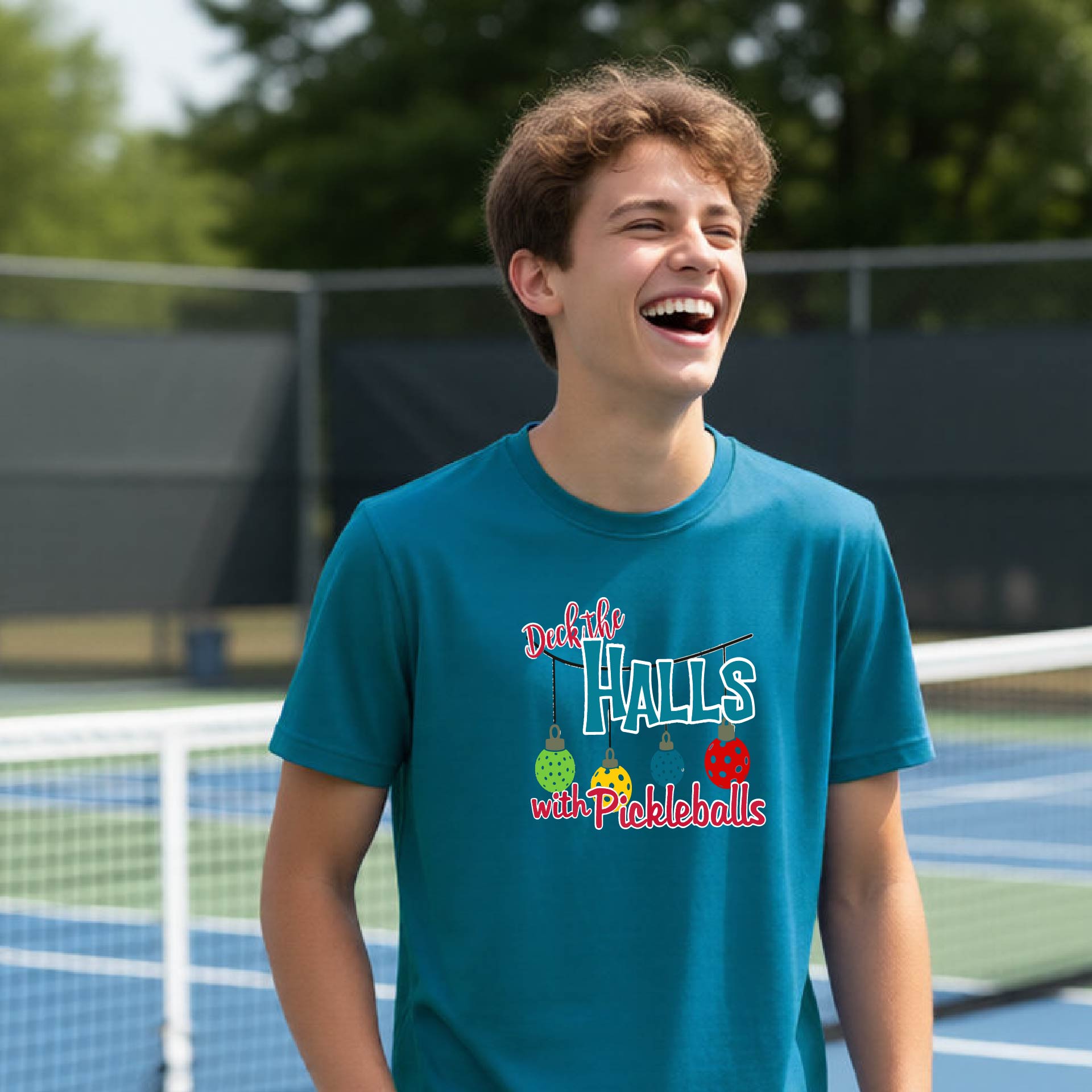A young man laughing on an outdoor pickleball  court wearing a teal graphic t-shirt that reads 'Deck the Halls with Pickleballs' with colorful pickleball graphics.