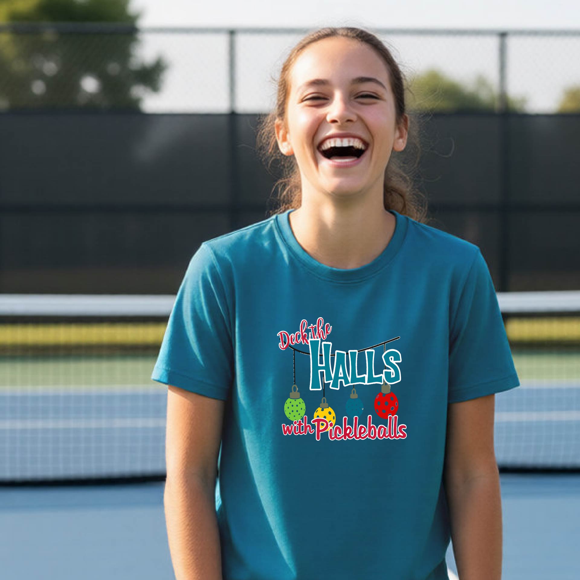 A young woman laughing on an outdoor pickleball court wearing a teal blue graphic t-shirt that reads 'Deck the Halls with Pickleballs' with a fishing rod and balls design.
