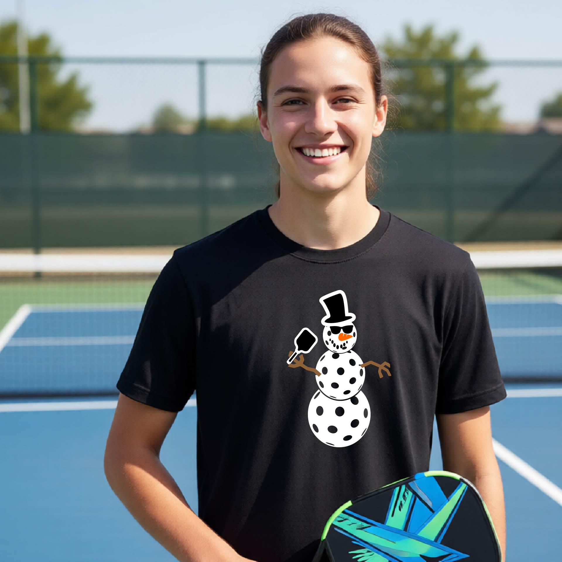 A smiling woman on a blue pickleball court wearing a black t-shirt with a white snowman pickleball graphic holding a paddle.