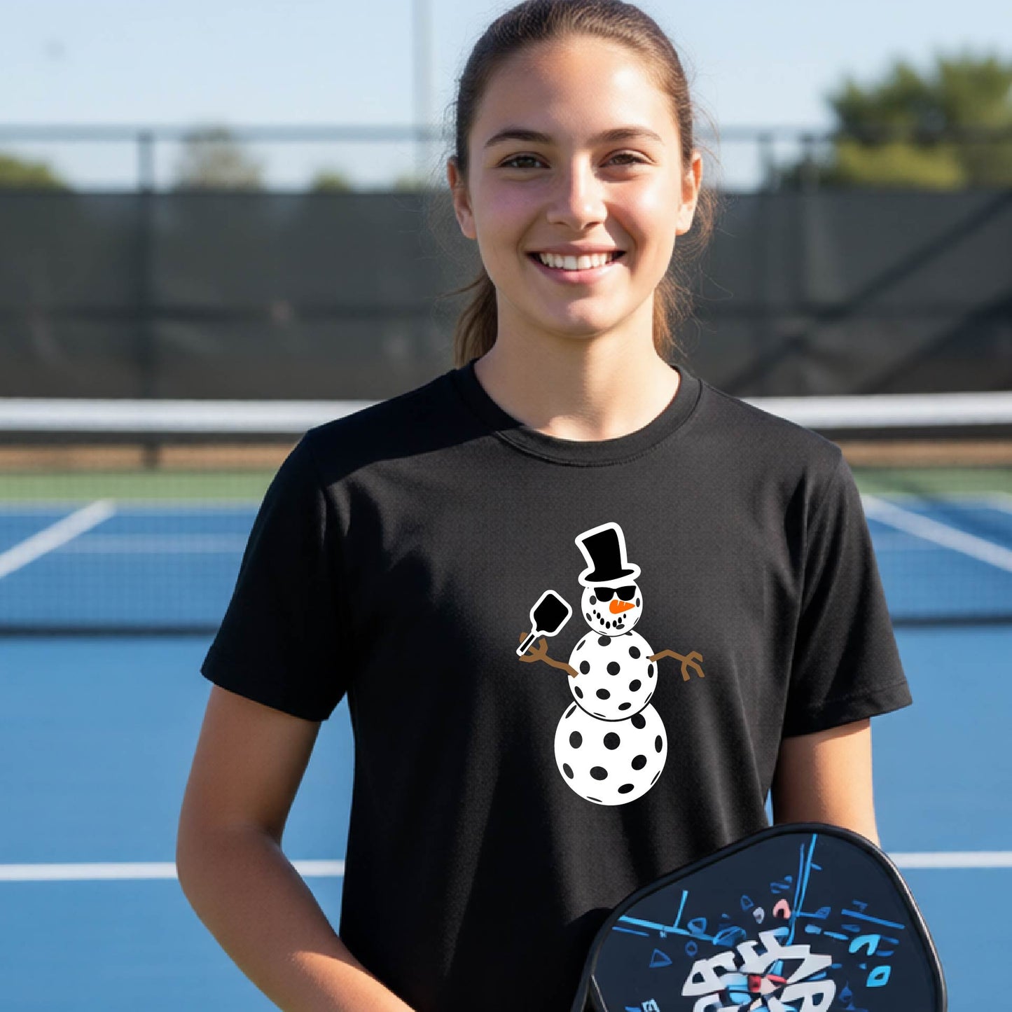 A smiling young woman stands on an outdoor pickleball court wearing a black t-shirt with a white snowman graphic holding a snow shovel. She is holding a black pickleball paddle in her hand.