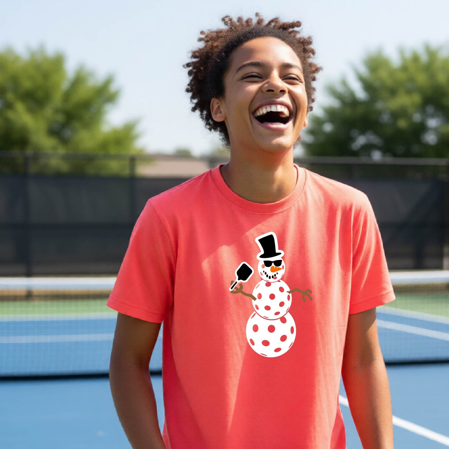 A smiling person standing on a blue pickleball court wearing a coral-pink short-sleeve T-shirt with a cartoon snowman pickleball graphic on the chest.