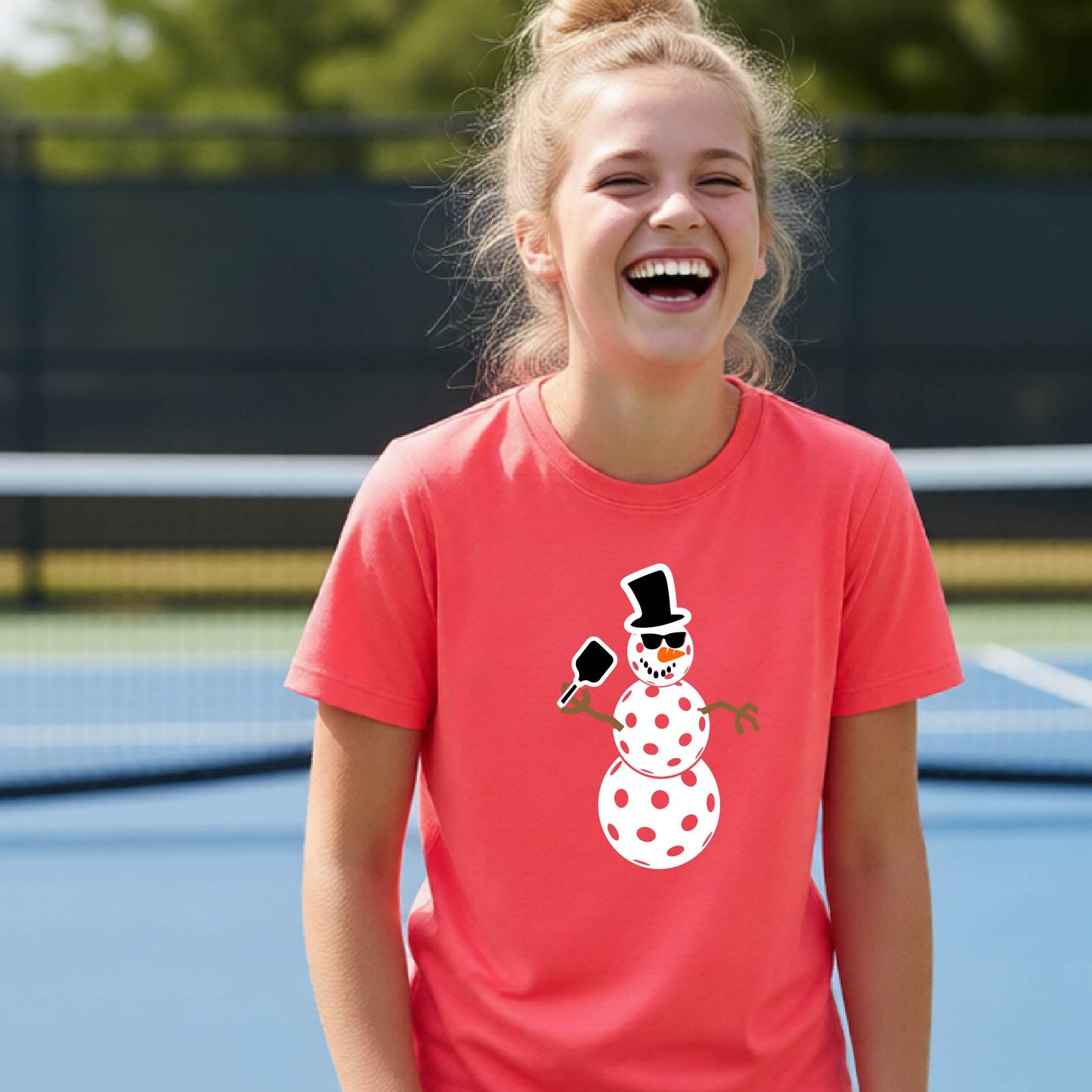 A young girl with blonde hair in a bun is laughing on a blue pickleball  court, wearing a coral pink t-shirt with a cartoon snowman pickleball graphic.