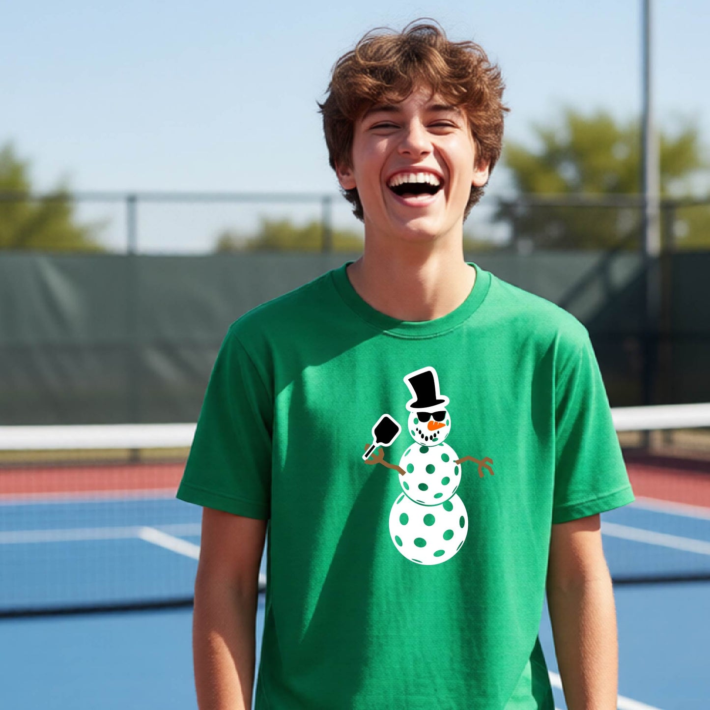A young man laughing on an outdoor pickleball  court wearing a bright green t-shirt with a cartoon snowman pickleball graphic on the front.