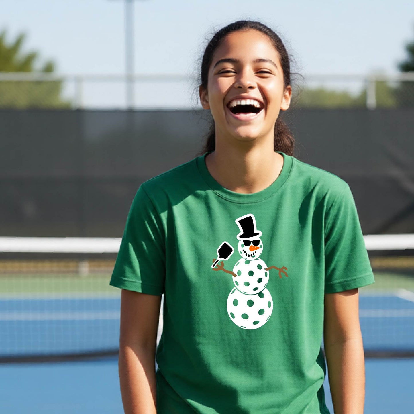 A woman laughing on a pickleball court wearing a green crewneck t-shirt with a cartoon snowman pickleball graphic on the chest.