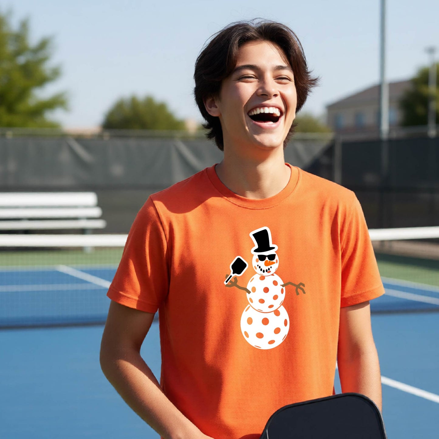 A young man laughing on an outdoor pickleball  court wearing a bright orange t-shirt with a cartoon snowman pickleball graphic on the front.